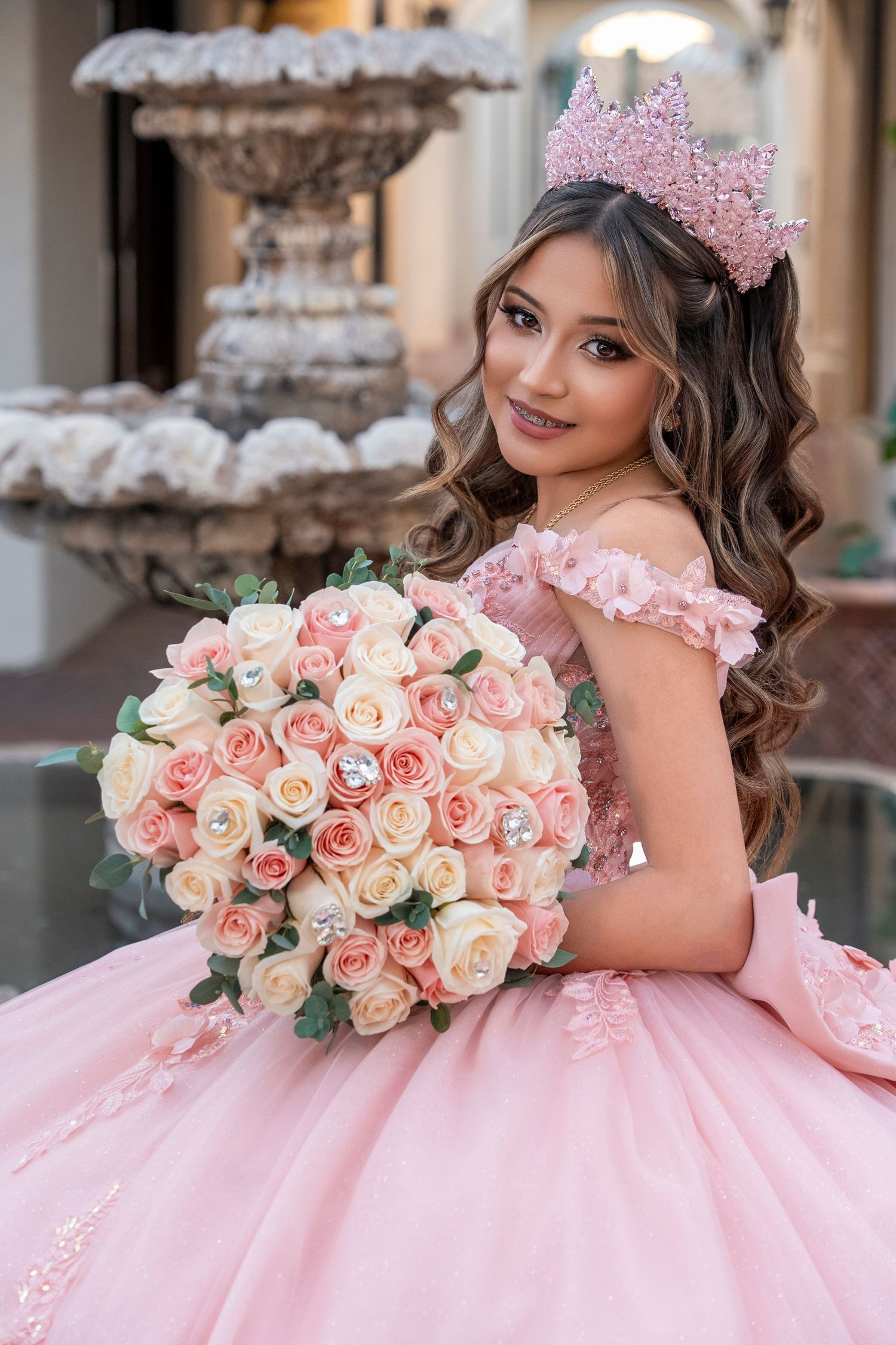 Young woman in pink quinceañera gown and crown holds large bouquet of pink and white roses near ornate fountain.