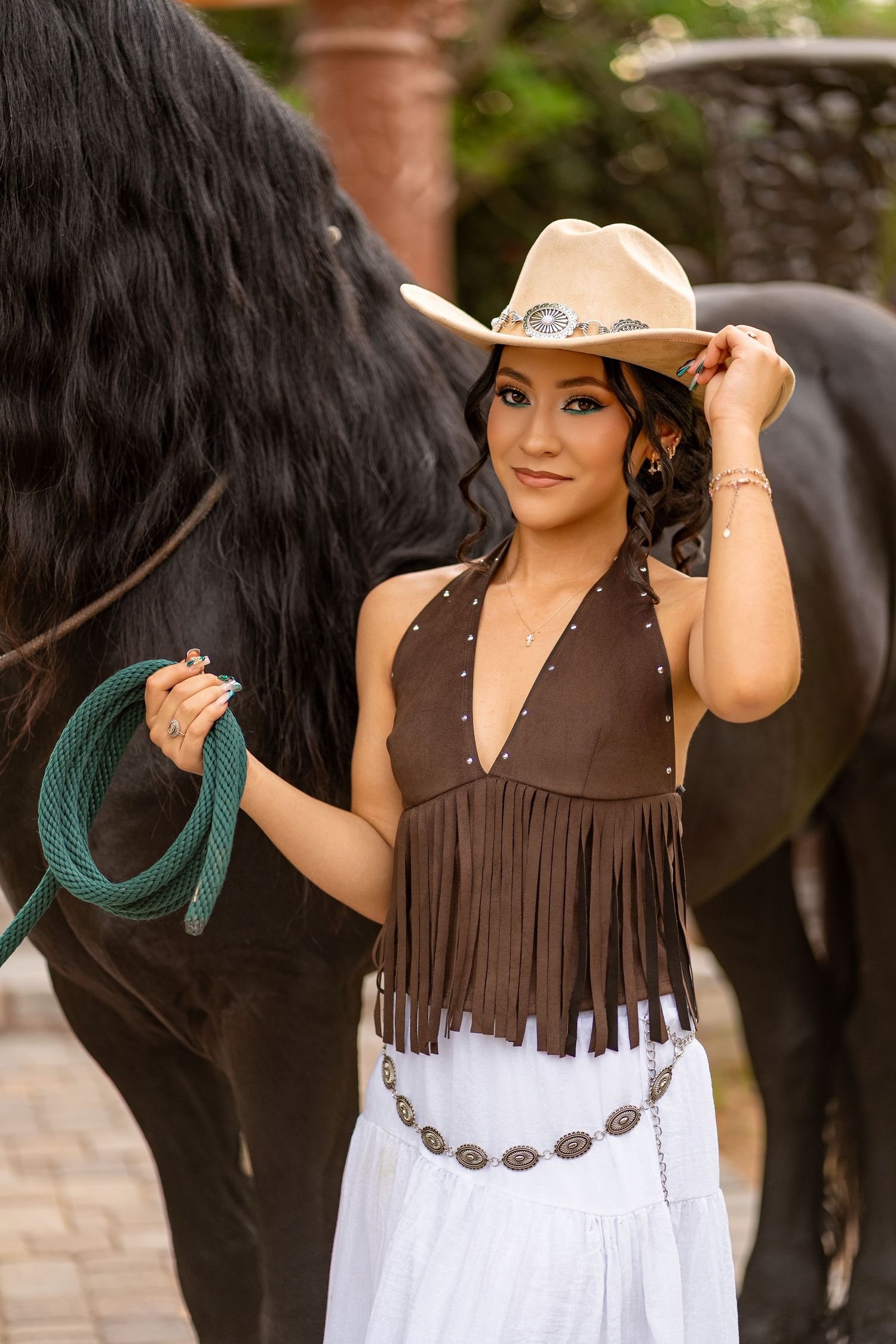 Young woman in cowboy hat and fringed western top holds horse lead rope while posing with black horse outdoors.