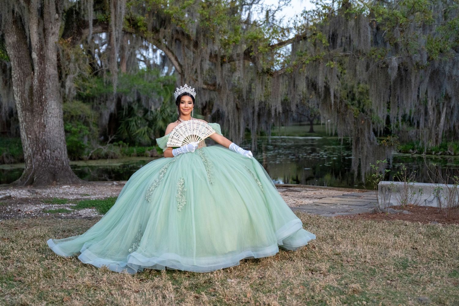 Young woman in elegant mint green quinceañera ball gown poses outdoors under Spanish moss-draped trees.