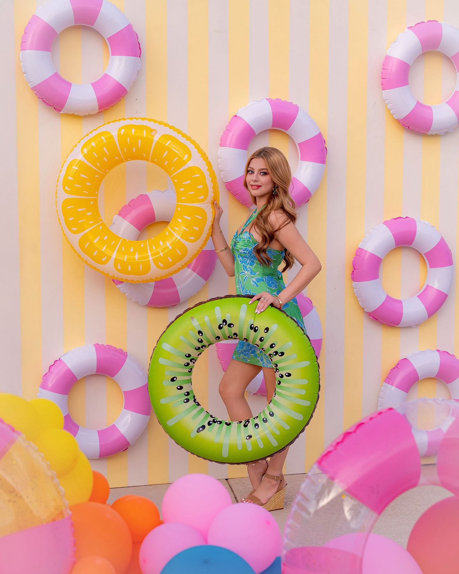 Woman in sequin swimsuit holding kiwi inflatable ring surrounded by colorful pool floats and pink striped wall.