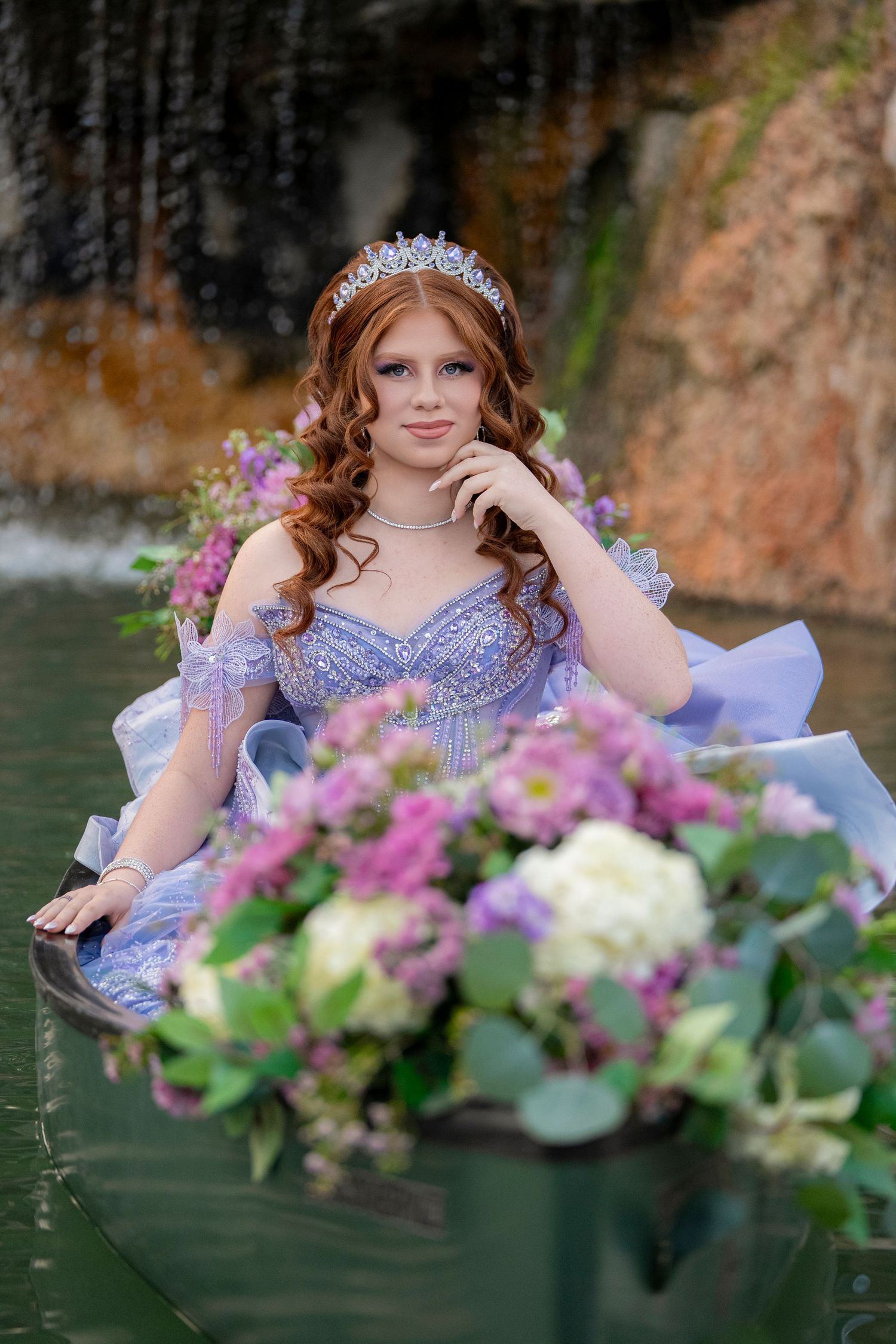 Red-haired girl in blue princess gown with tiara sits in flower-filled boat near waterfall.