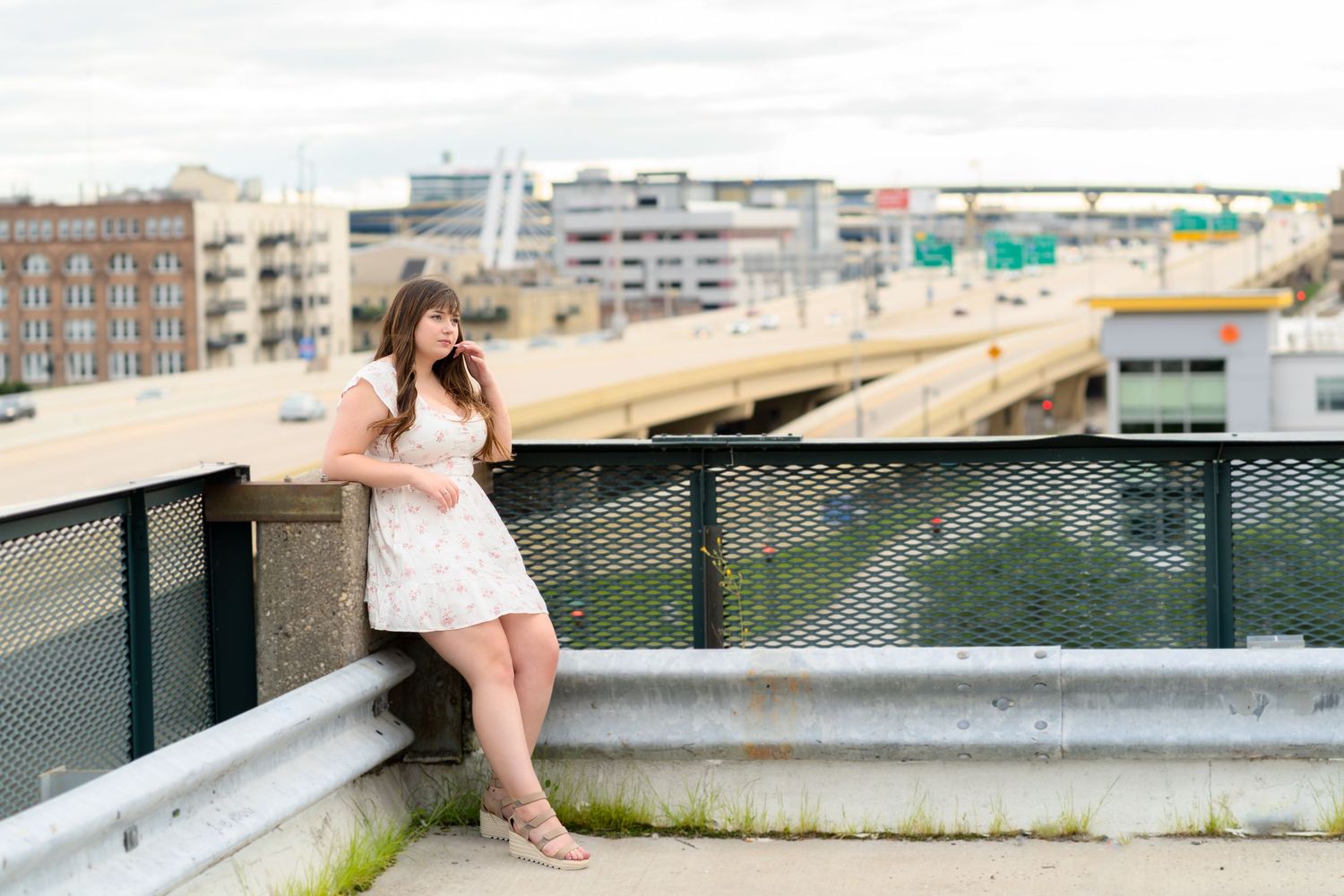 Someone in a white summer dress stands on an elevated deck overlooking a waterfront and highway in an urban setting.