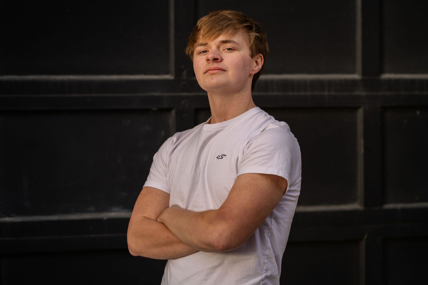 Someone in a white t-shirt stands confidently with arms crossed against a dark background in an outdoor setting.
