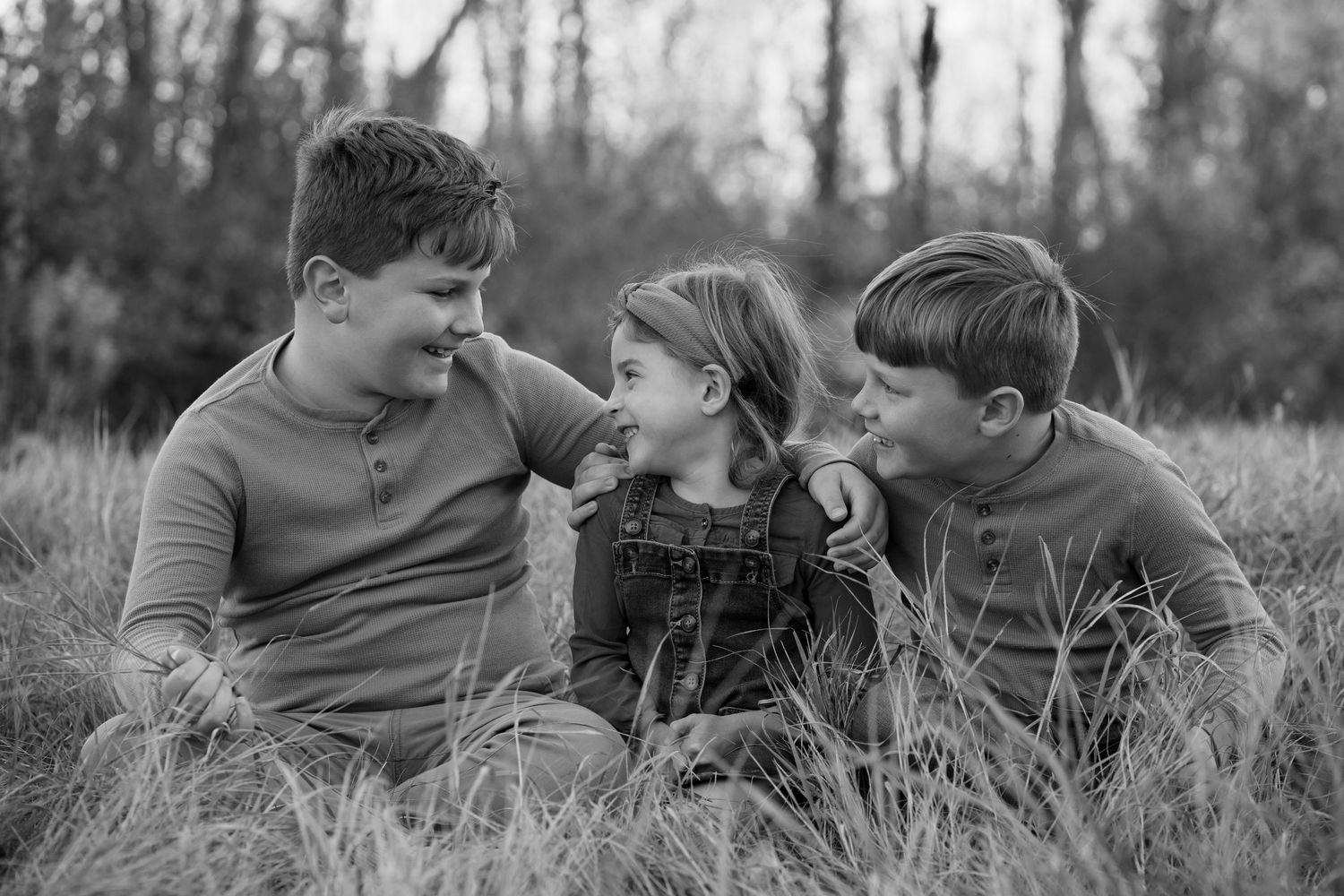 Black and white portrait of three children sitting in tall grass and looking at each other outdoors.