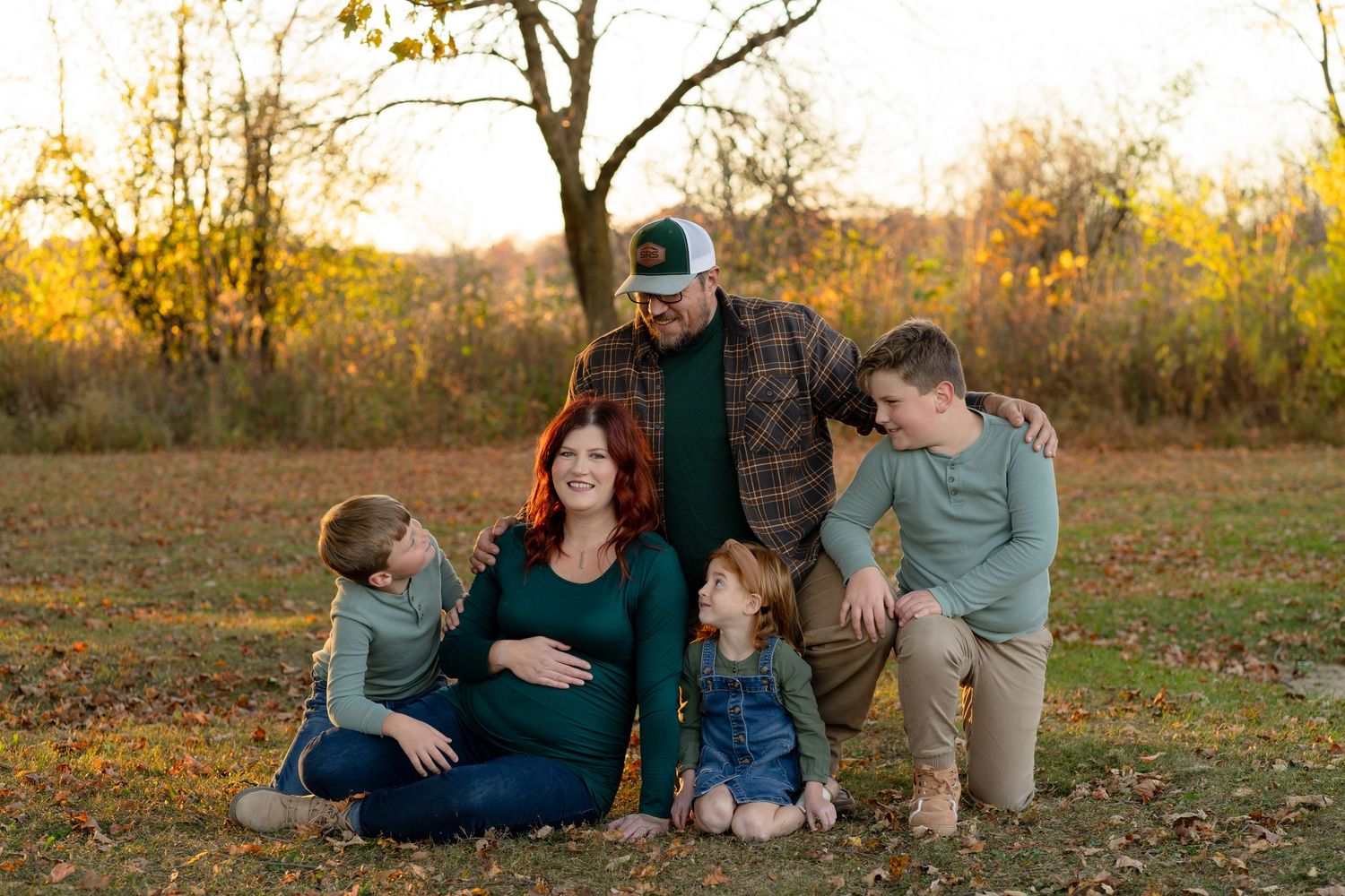 A pregnant woman in green sits on autumn ground with family members around her during a golden sunset.