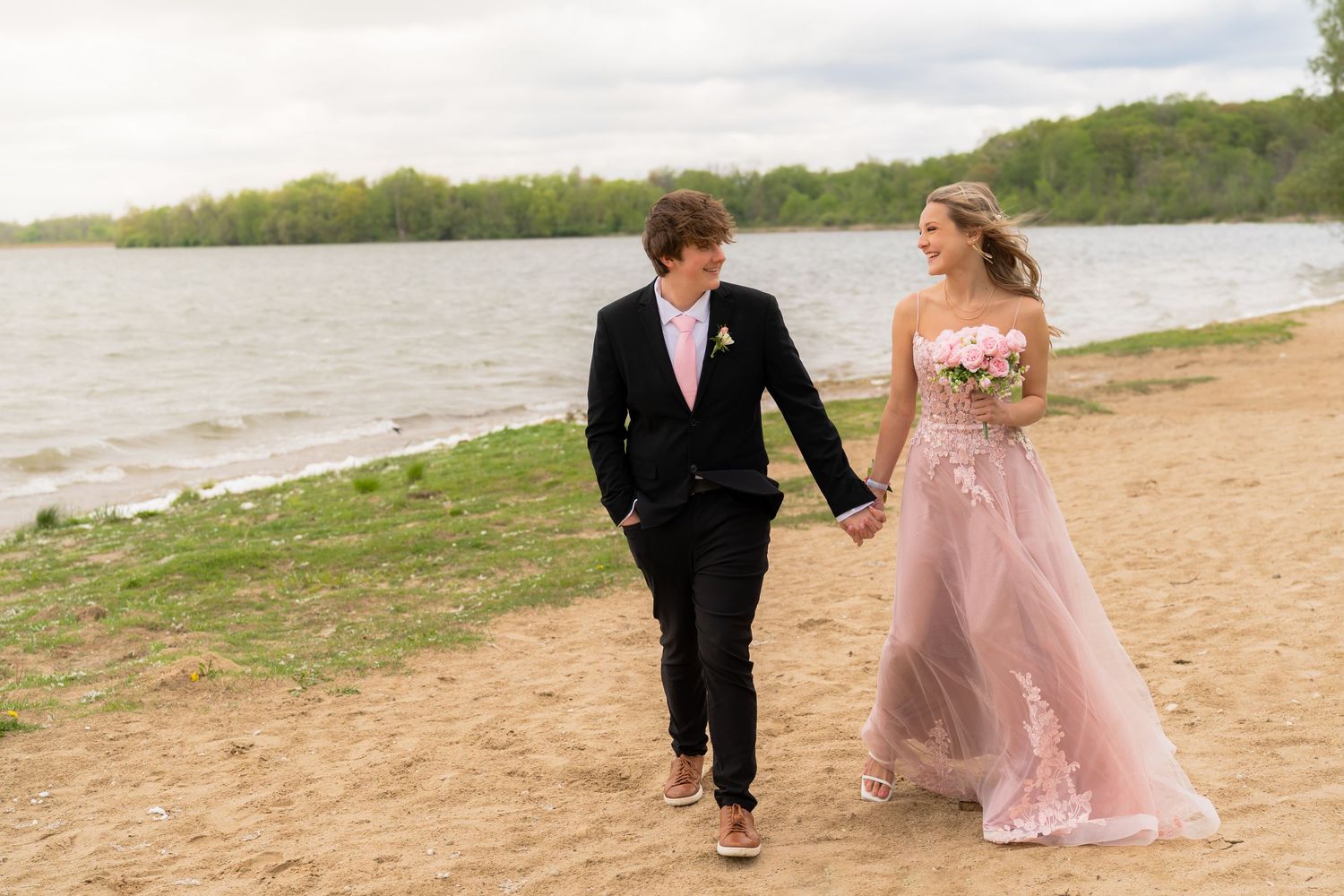 A couple in formal attire walks hand in hand along a sandy beach with trees and lake in the background.