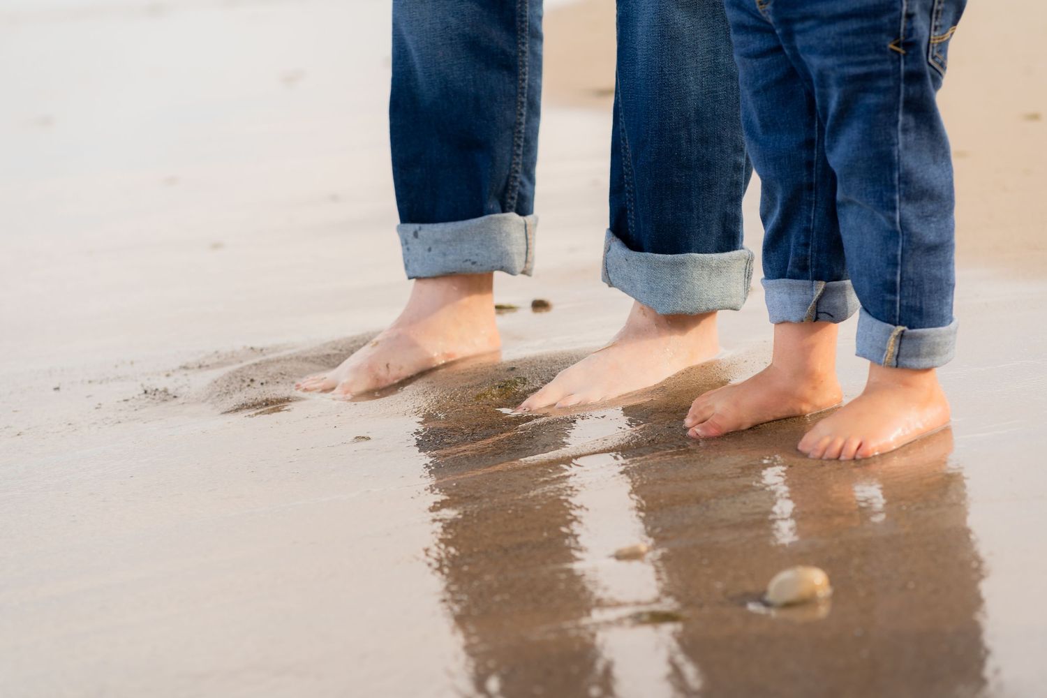 Bare feet and rolled up jeans walking along the ocean shoreline at sunset, leaving footprints in the wet sand.