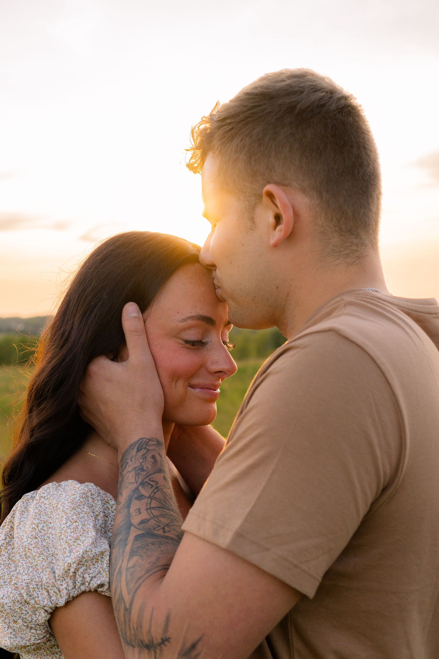 A romantic couple embraces during golden hour in a nature setting, with sun flare highlighting their intimate moment.