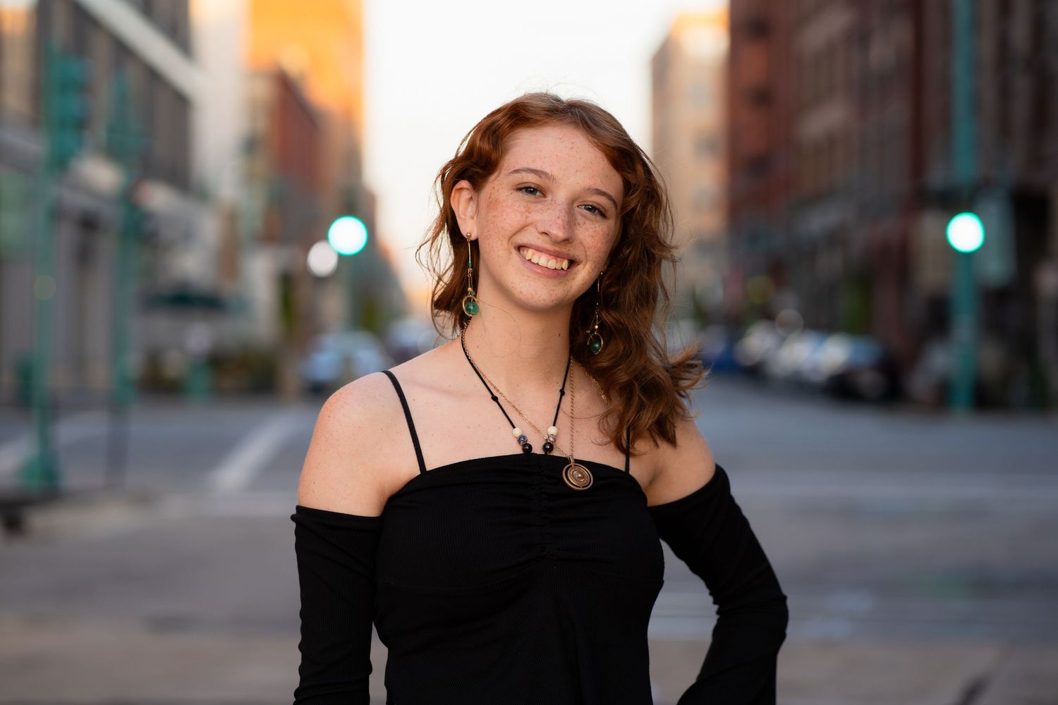 A woman in a black off-shoulder top smiles warmly on an urban street at sunset.