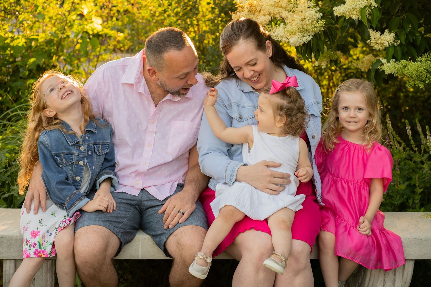 A cheerful group sits together on a bench outdoors surrounded by yellow flowers and greenery on a sunny day.