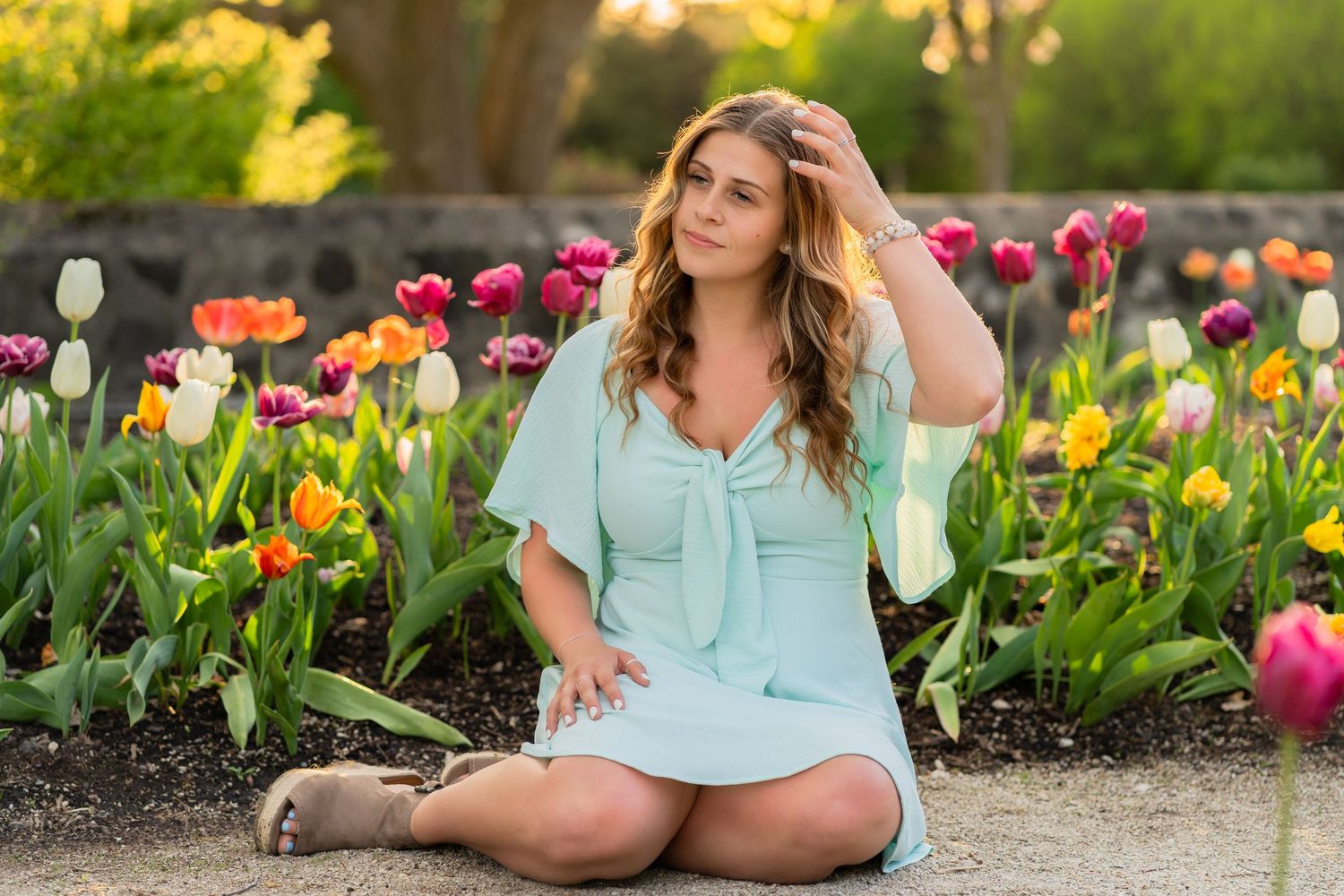 Woman in mint dress sits in garden among colorful spring tulips during golden hour sunset.