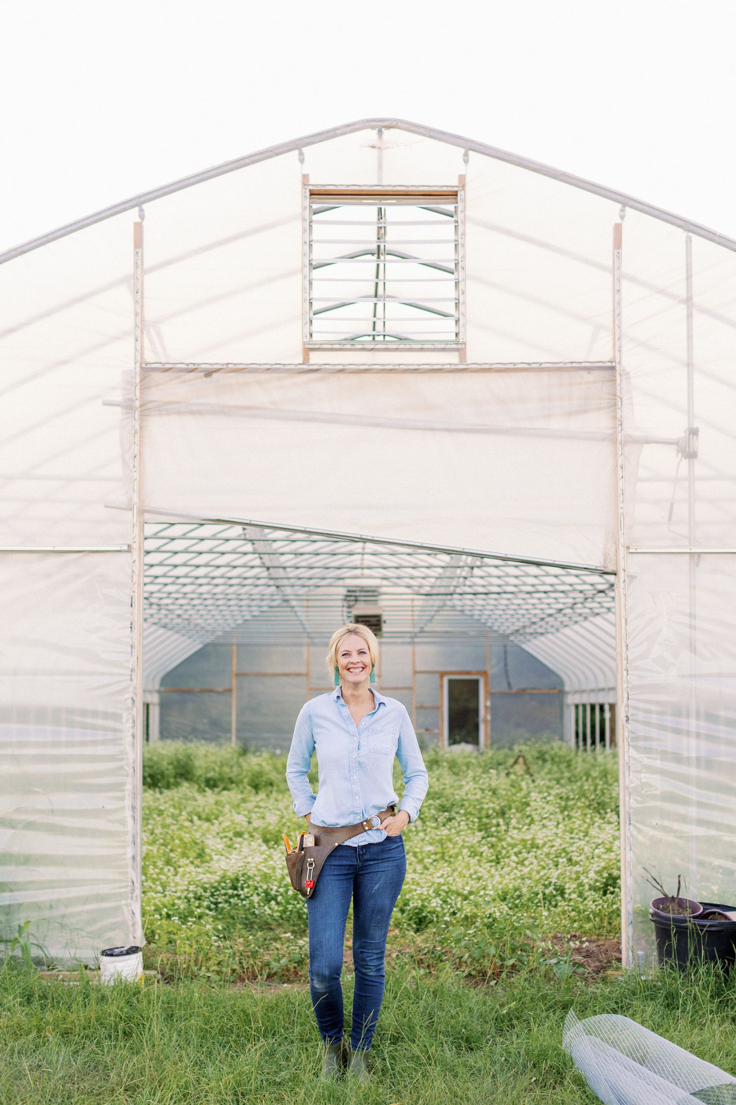 A farmer stands at the entrance of a greenhouse with white covering and metal framing structure in a rural setting.