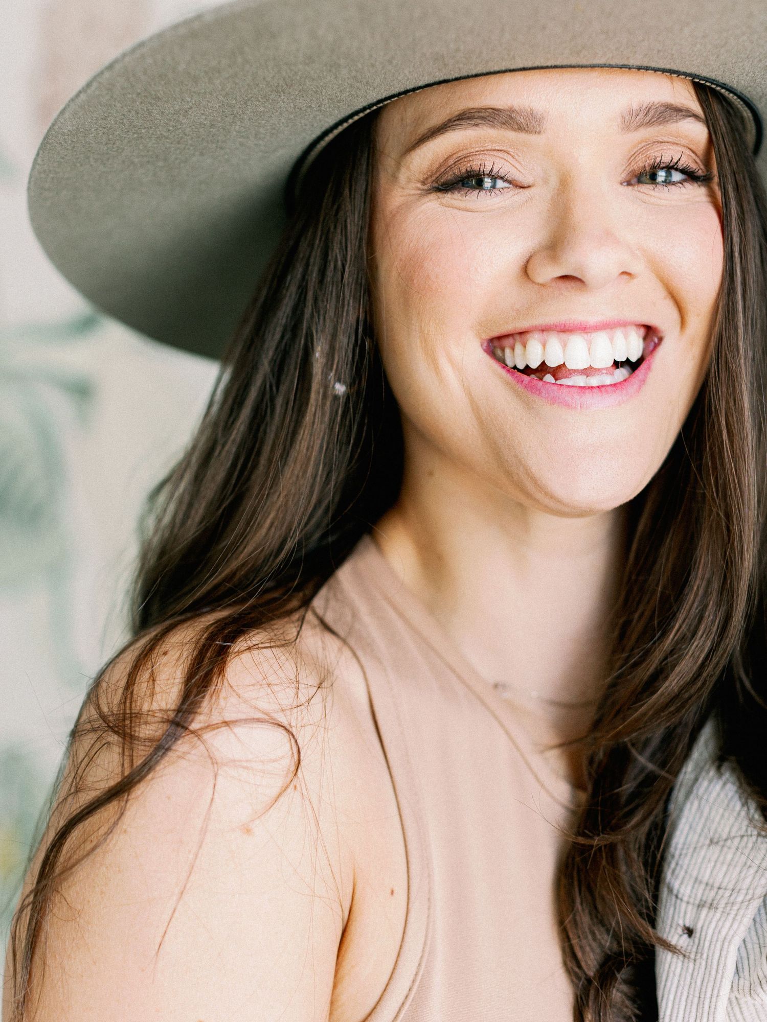 Smiling individual wearing a gray wide-brimmed hat and blush colored top against a light background.