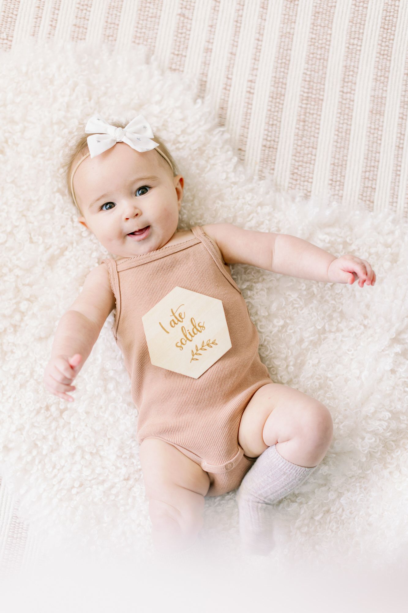 Baby wearing peach-colored romper with bow headband lying on white textured blanket.