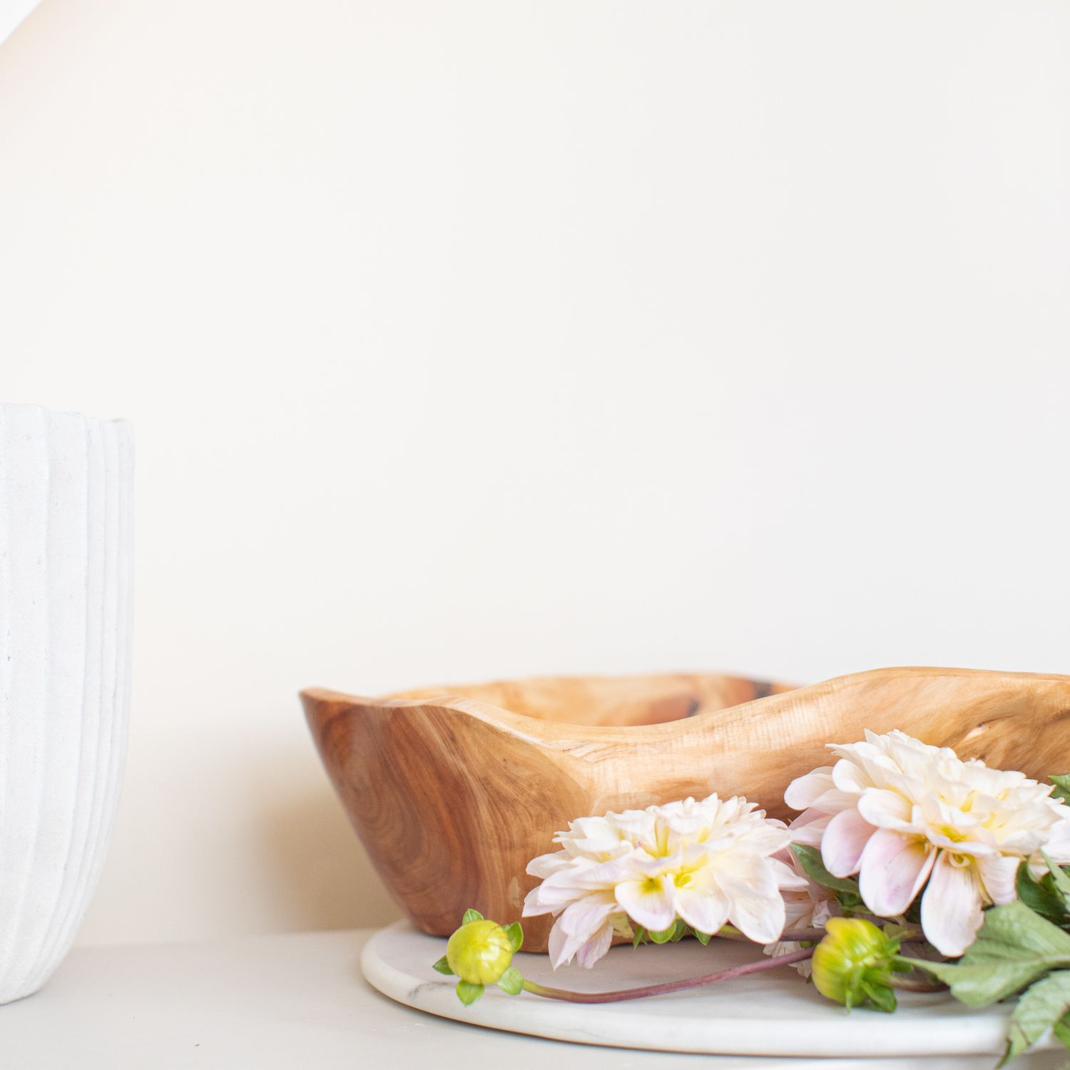 Wooden bowl with delicate pink dahlias sits beside a white glass on a light minimalist surface.