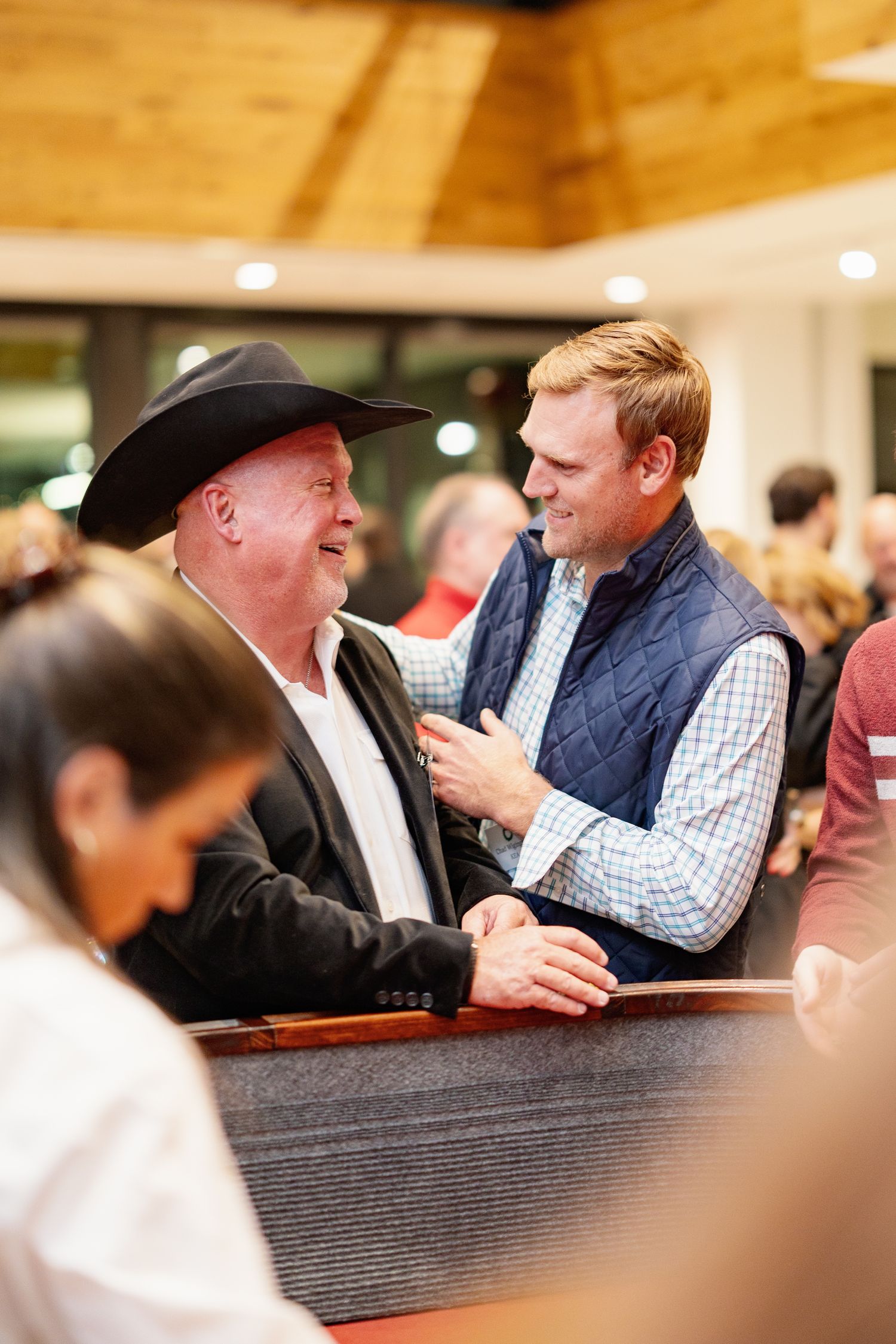 Two men engaged in friendly conversation at an indoor event, one wearing a cowboy hat and one in a navy vest.