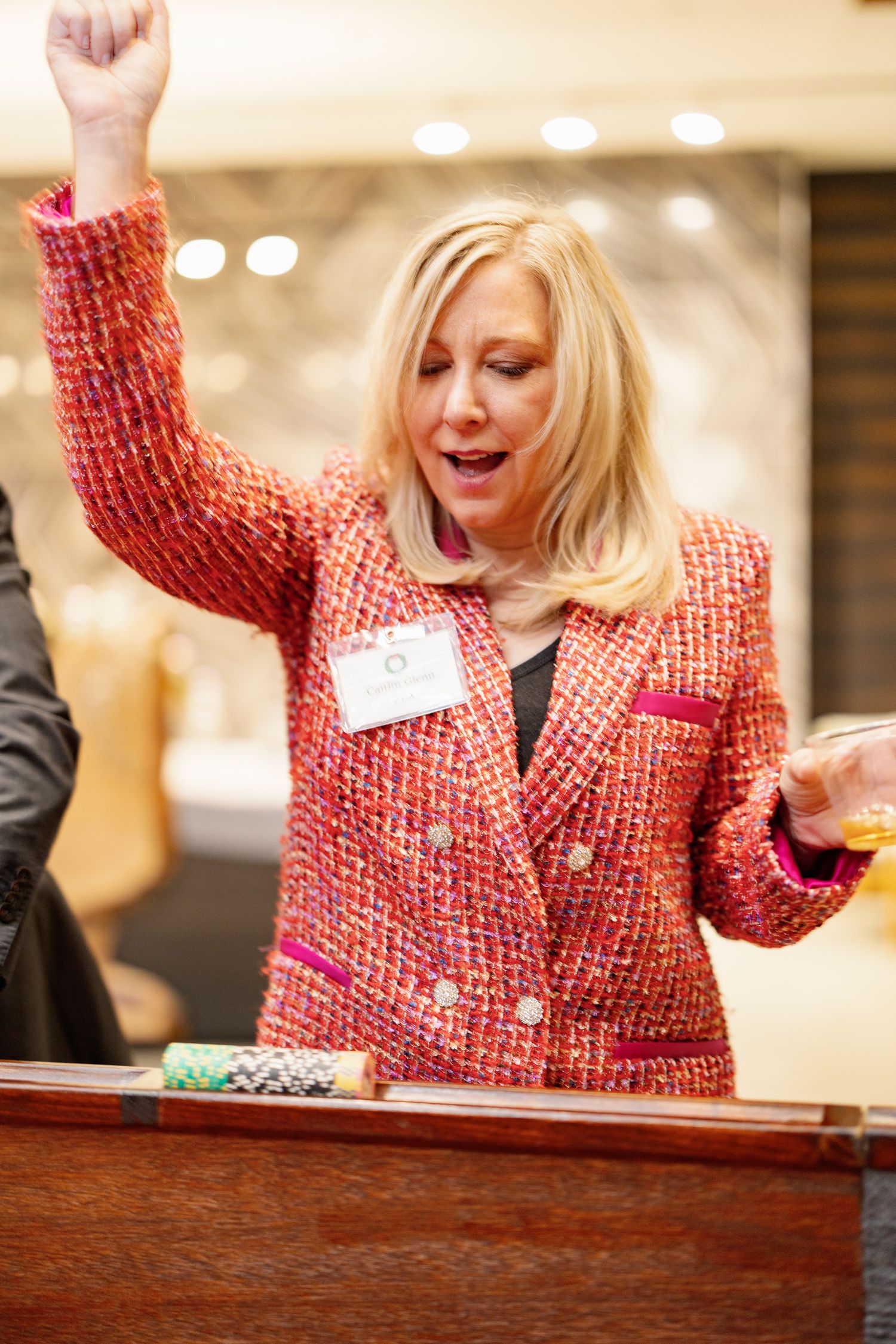Woman in red tweed blazer celebrates enthusiastically with raised fist at event with name tag visible.