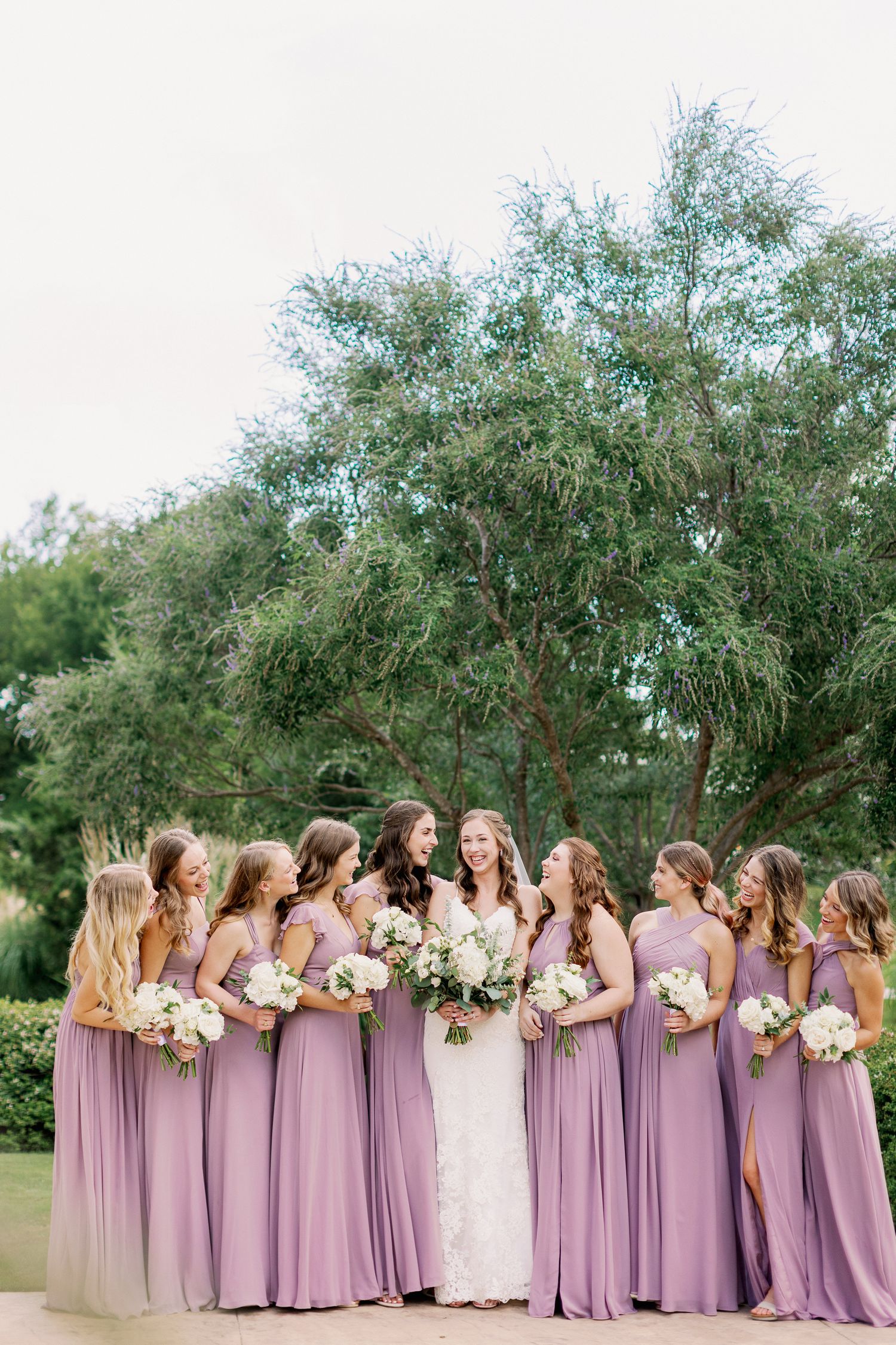 Bridesmaids in flowing lavender dresses stand together with bouquets against a backdrop of lush greenery.