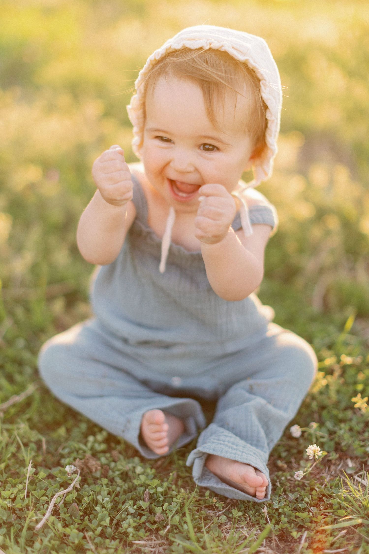 Small child wearing blue overalls and white bonnet sits in grass during golden hour with joyful expression.