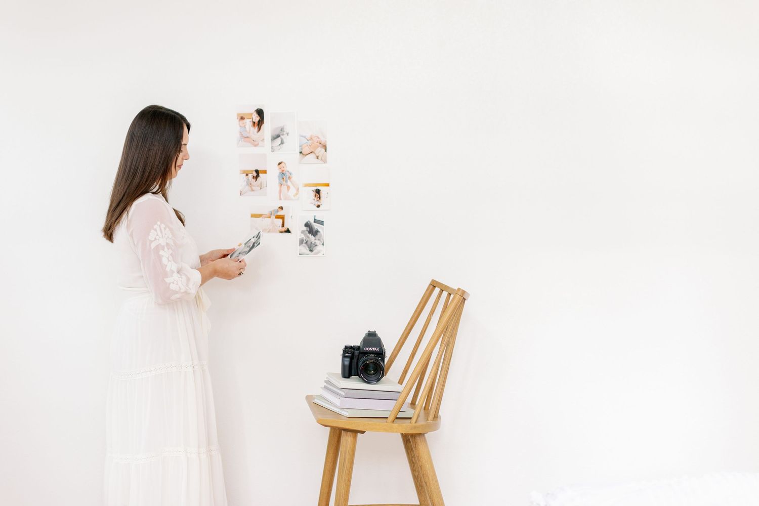 Photographer in white dress examines photo prints on white wall next to wooden chair with camera.