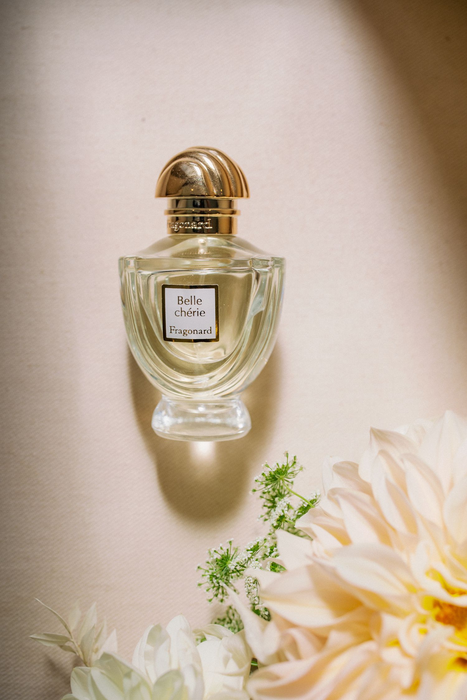 An elegant glass perfume bottle with gold cap sits beside delicate peach-colored flowers and white floral accents.