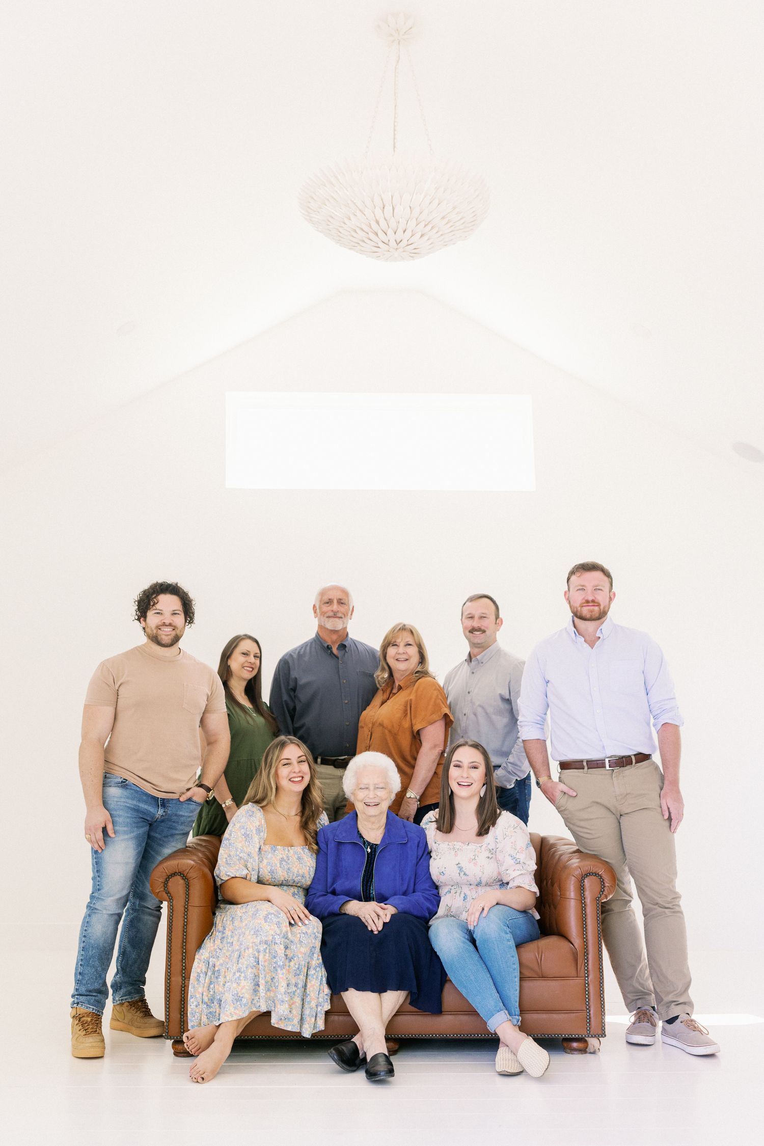 Multi-generation family portrait in a minimalist white studio setting with a brown leather couch and pendant light.