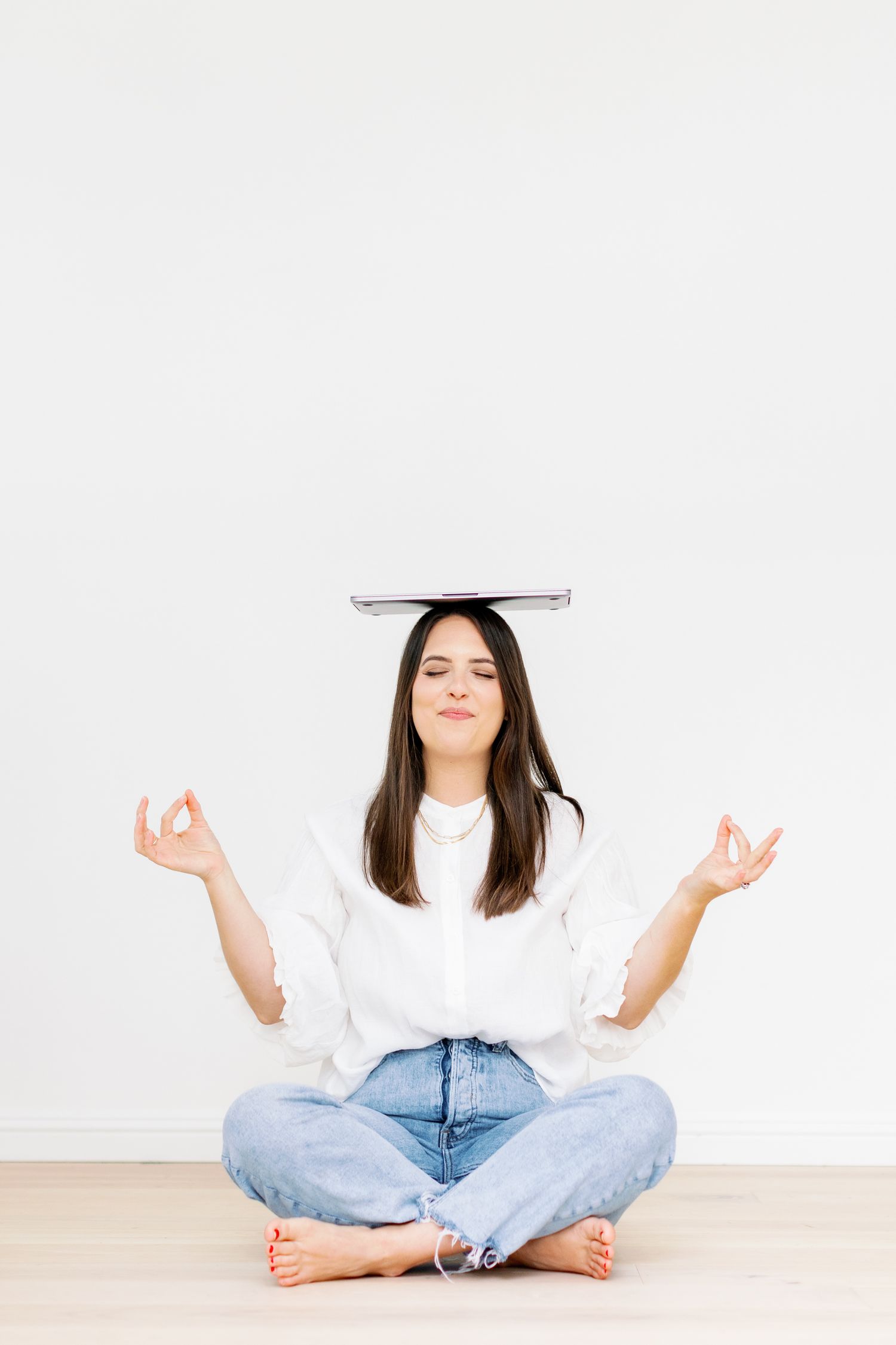 Person in white shirt and jeans sitting cross-legged on floor with book balanced on head in meditation pose.