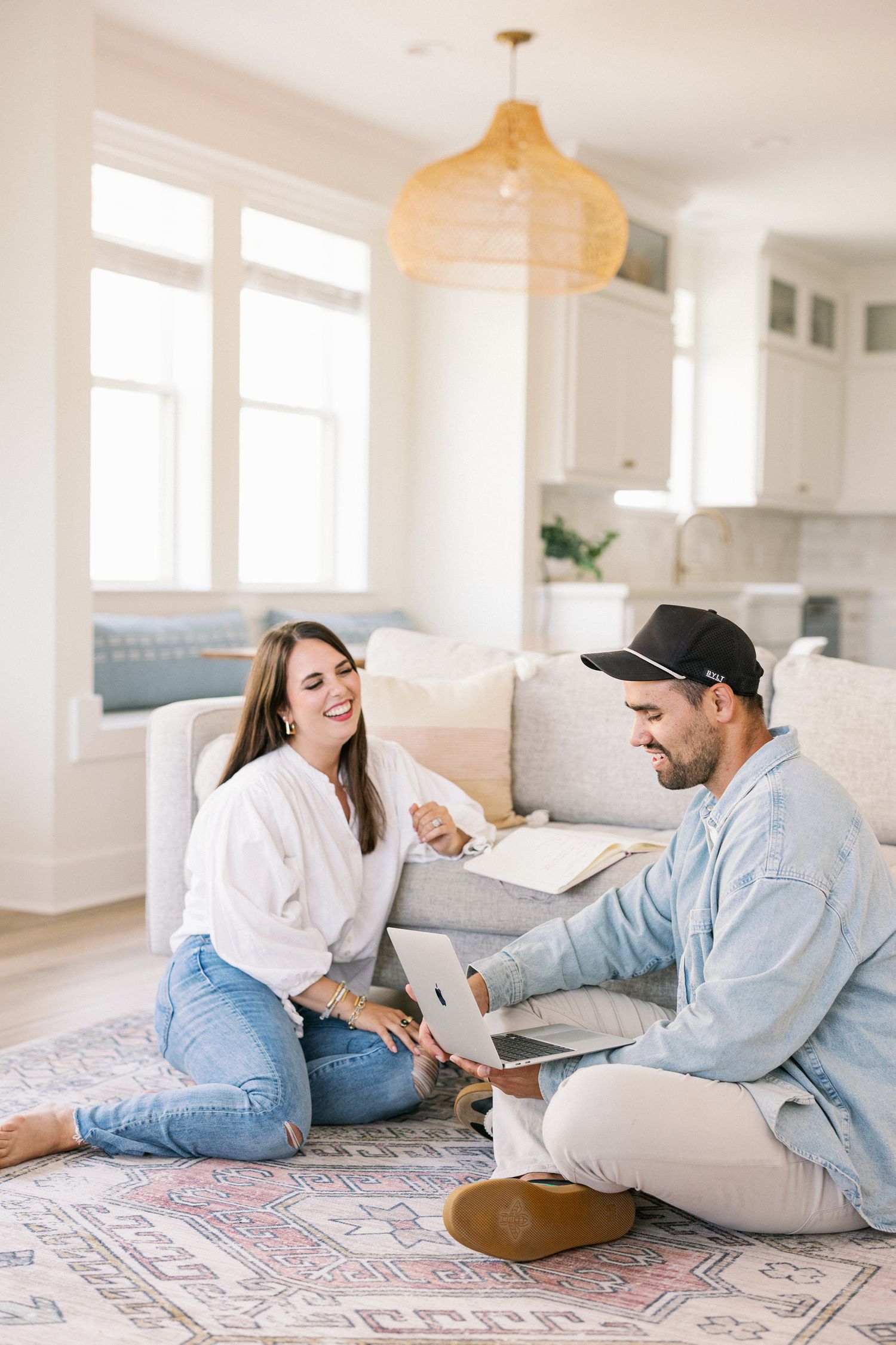 Two people sit on a patterned rug in a bright living room with white kitchen cabinets and modern decor.