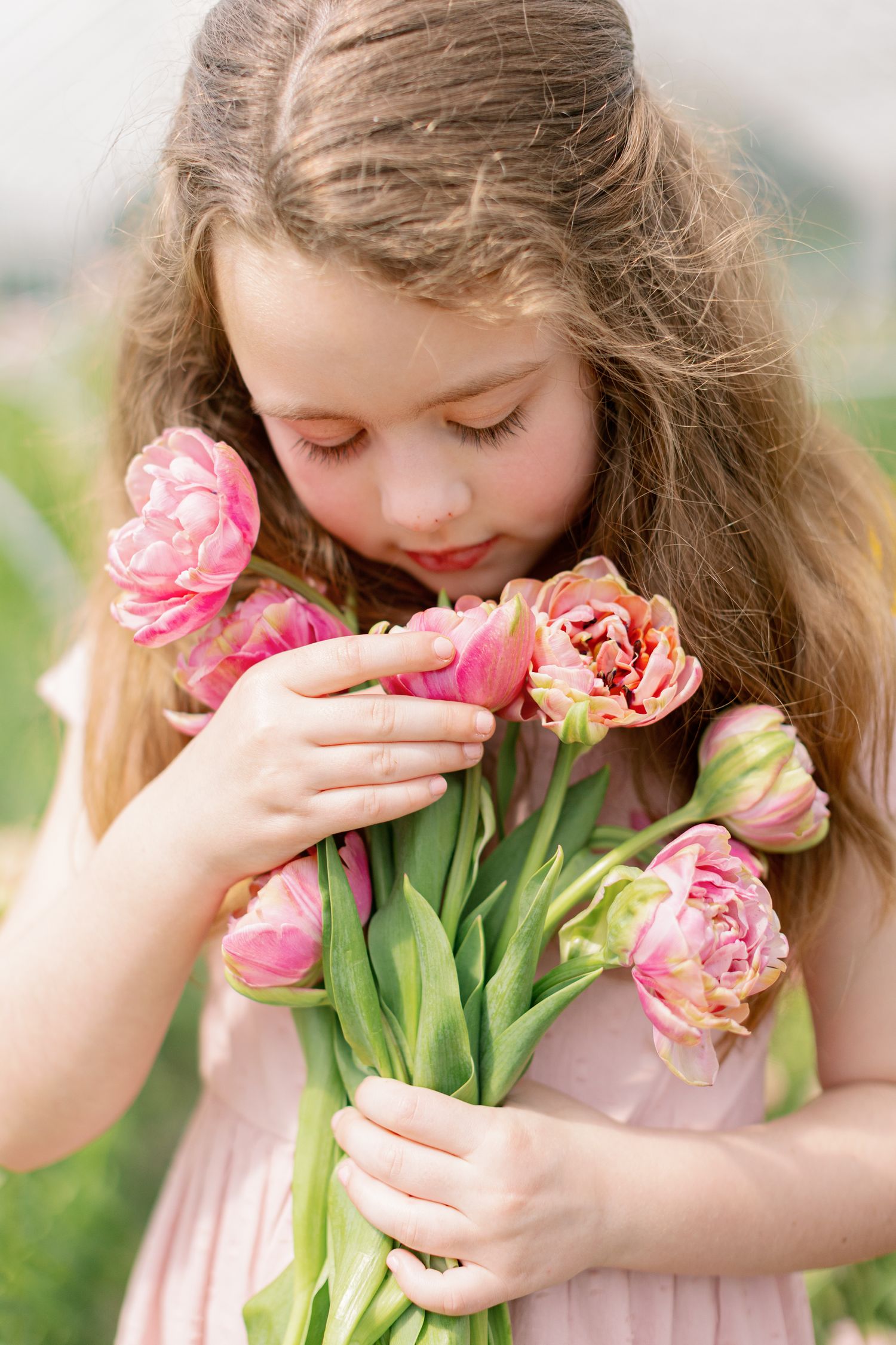 Young girl in pink dress smells bouquet of pink peony tulips while holding them close in outdoor garden setting.