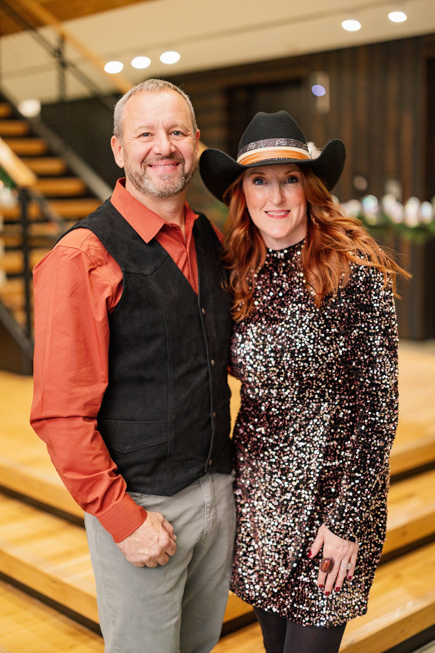 A couple dressed in western style attire pose on wooden stairs with a man in orange shirt and a woman in sequin dress.
