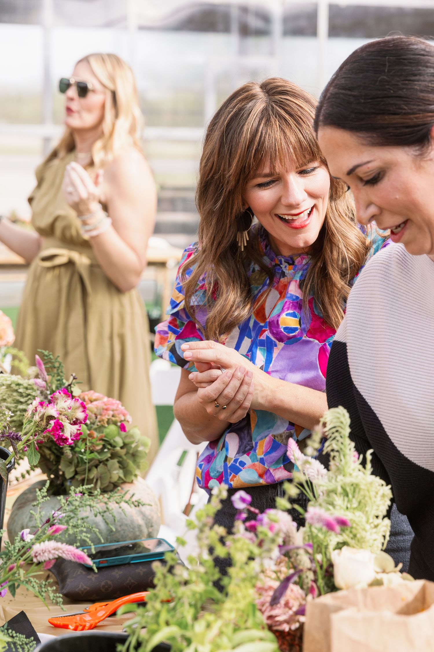 Ladies gather around fresh produce and flowers at an outdoor market while sharing laughs and enjoying a sunny day.