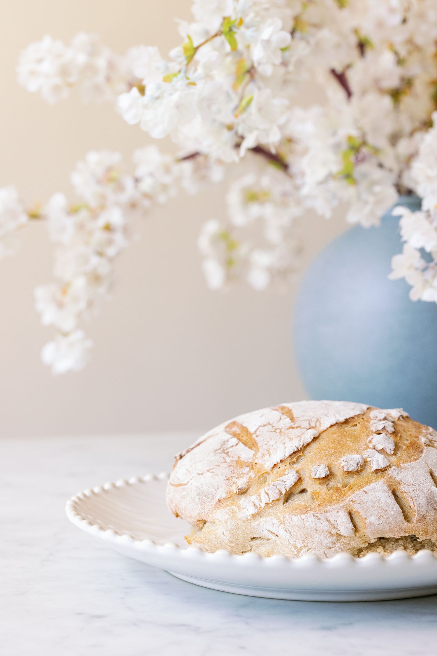 Fresh homemade crusty bread on white plate with spring cherry blossoms in blue vase against light background.