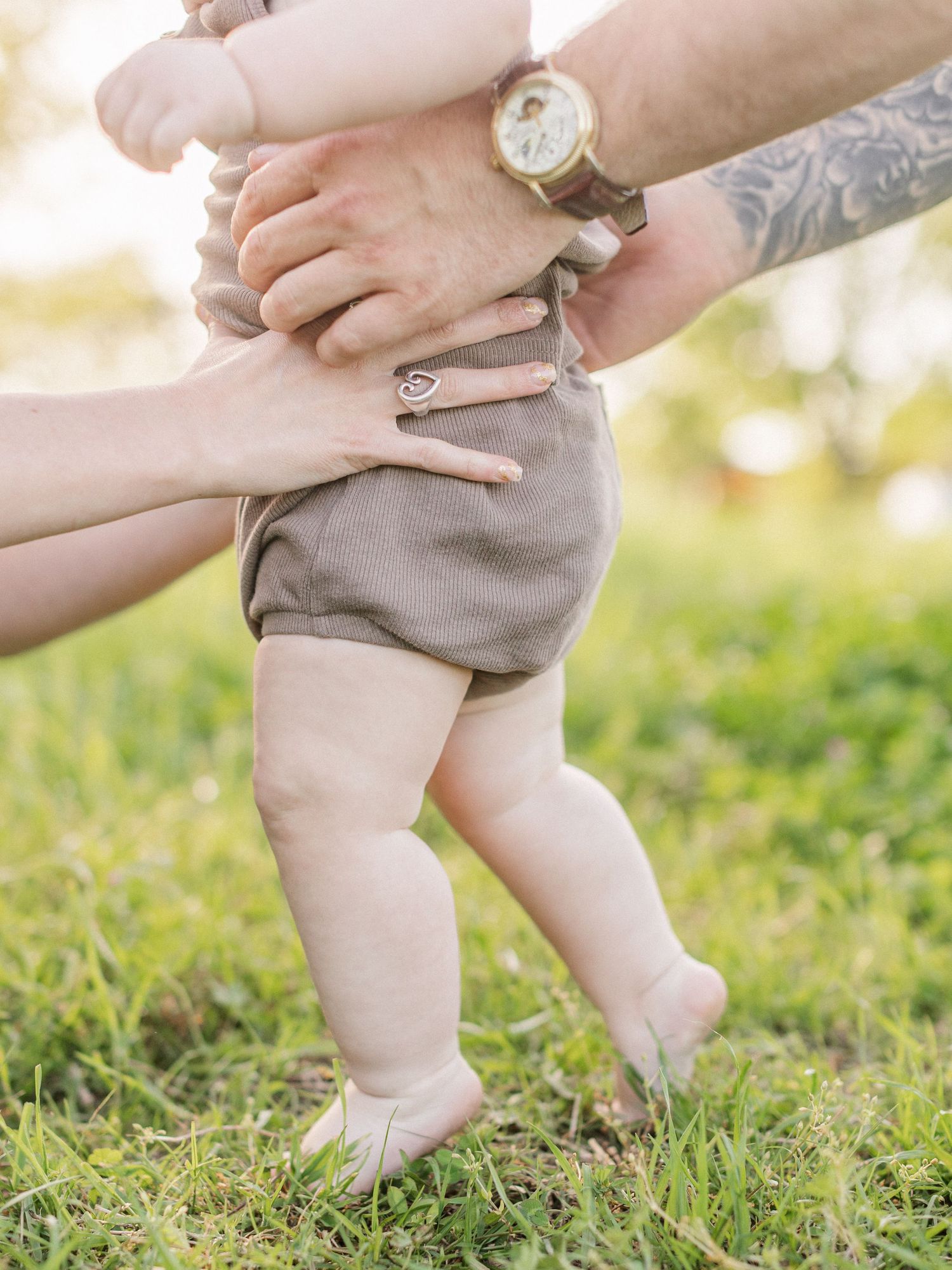 Parent's tattooed hands holding baby's feet in grey shorts standing on grass in warm sunlight.