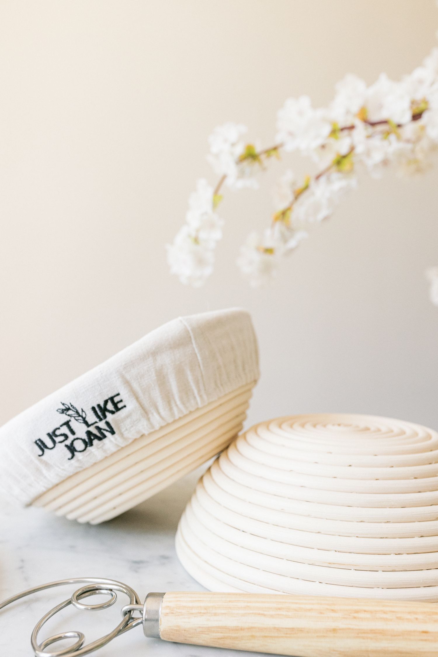 A woven bread proofing basket with white ceramic bowl and kitchen utensils against a light background with cherry blossoms.