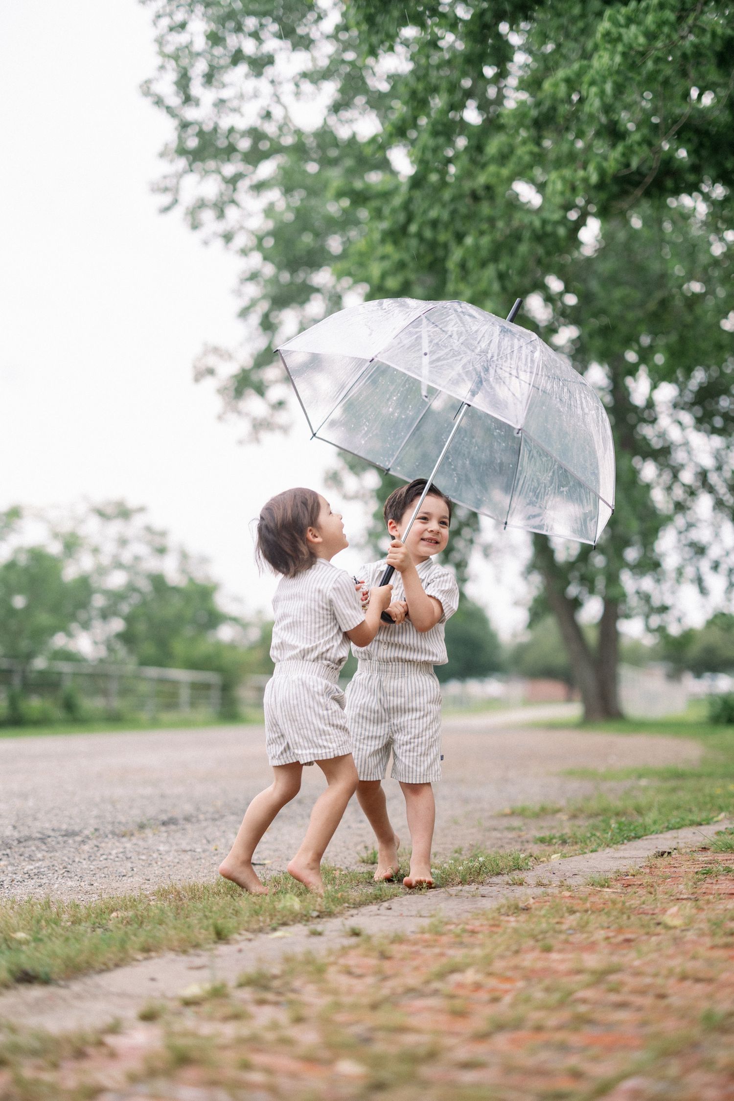 Two children playing together under a clear umbrella on a tree-lined path during a cloudy day.