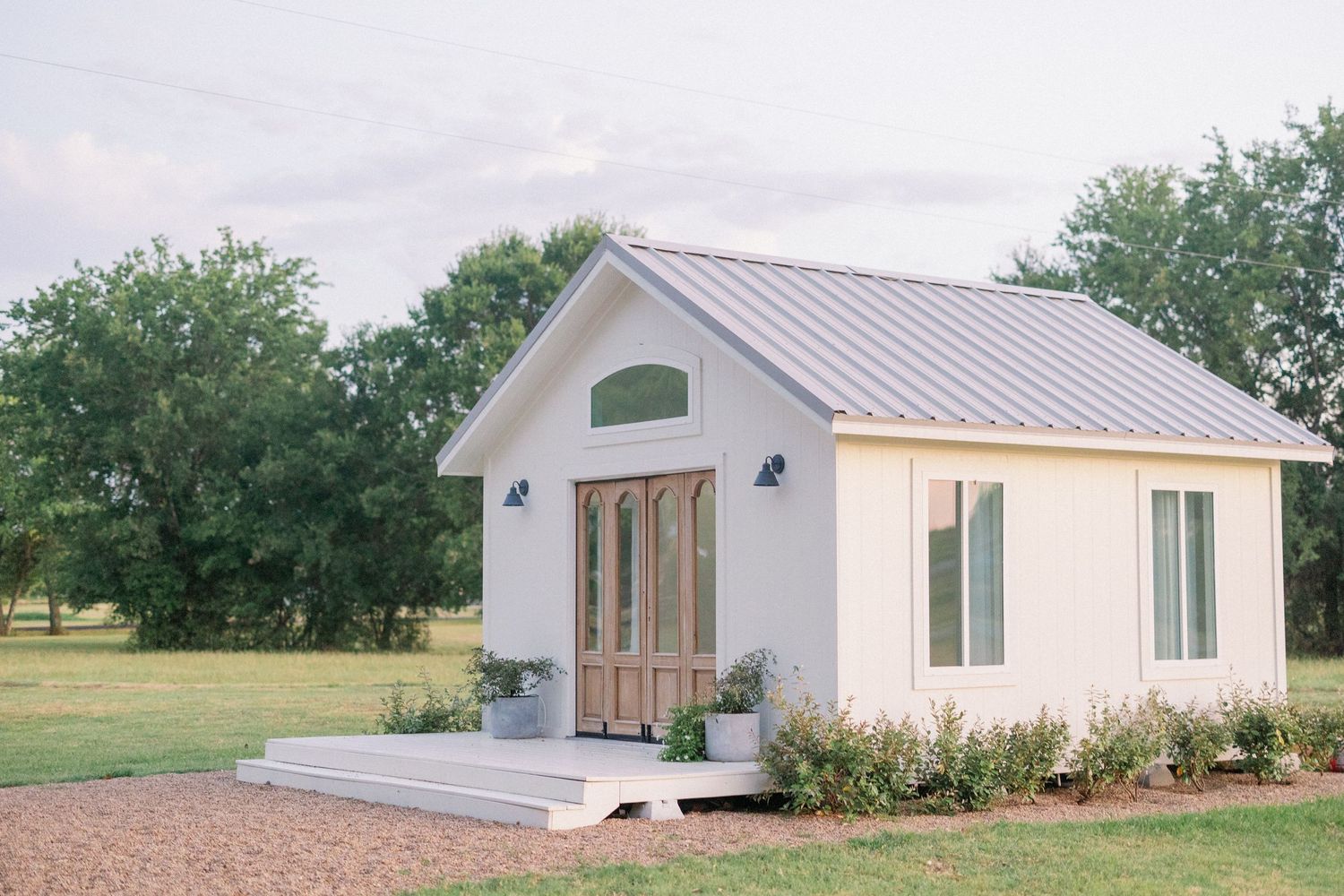 Small white modern tiny house with metal roof and wooden doors sits on platform with landscaped garden border.