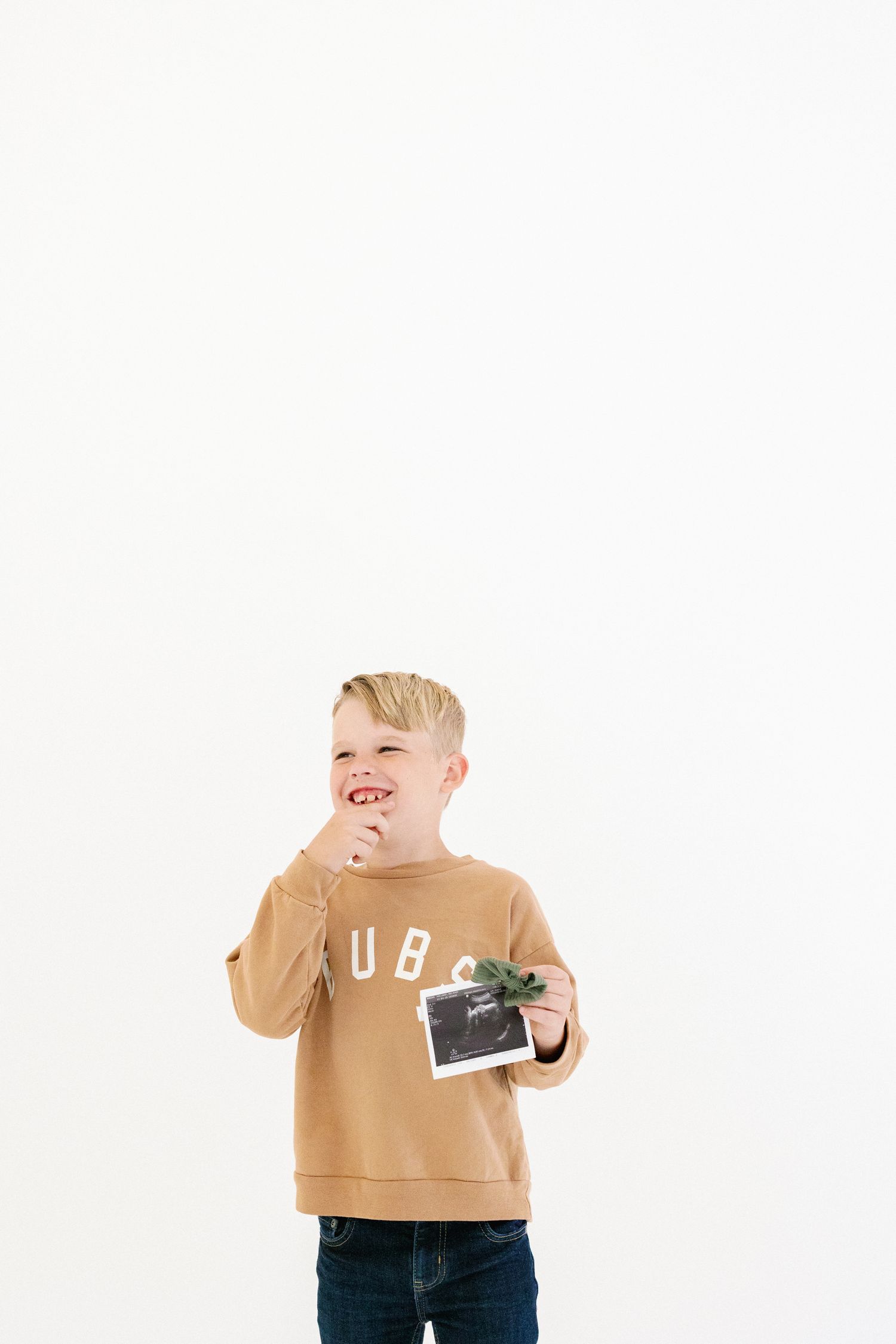 Child in a tan sweatshirt holds an ultrasound photo while standing against a white wall.
