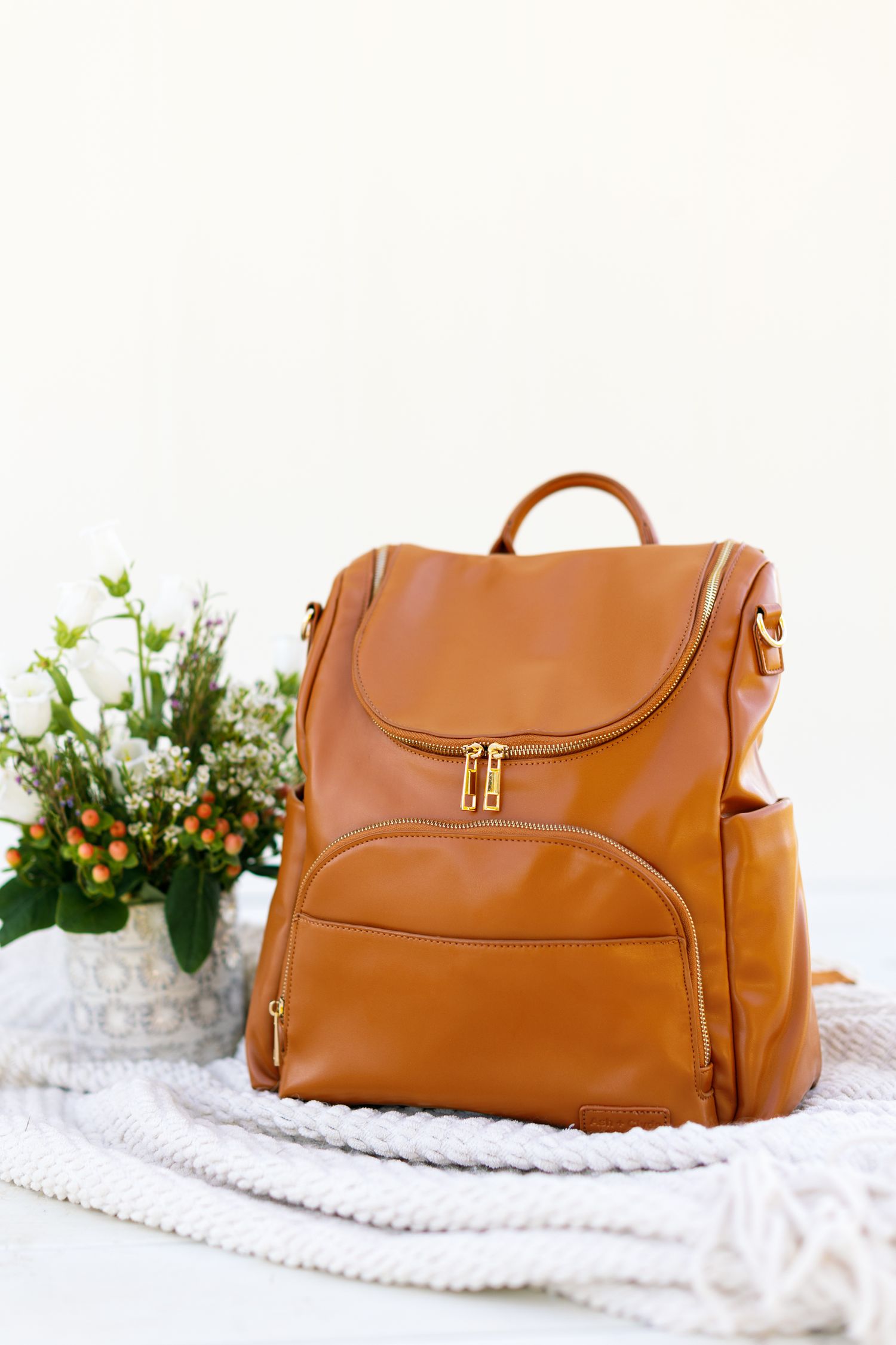 Brown leather backpack diaper bag with flap closure and pockets sits beside white flowers on a cream blanket.
