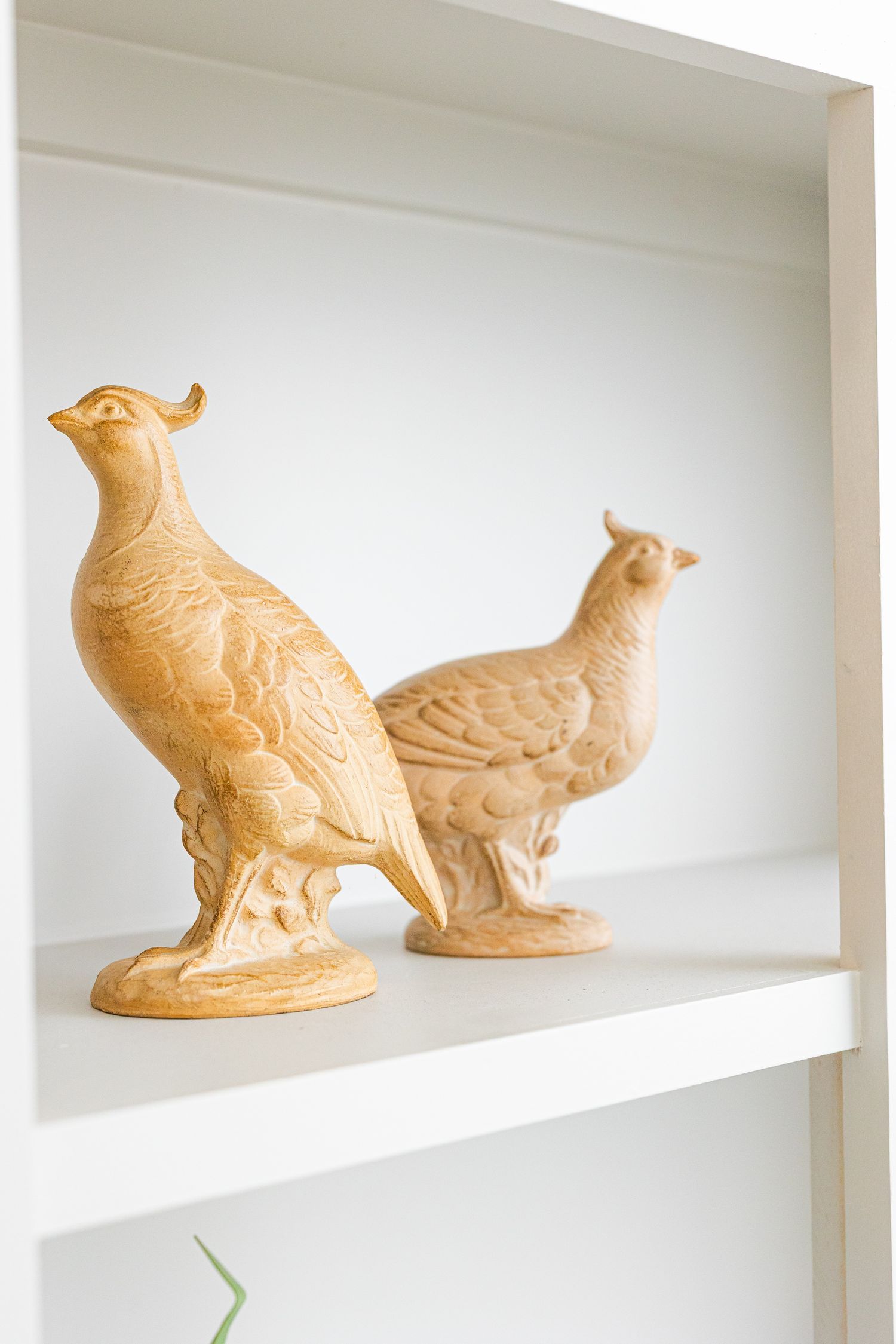 Two decorative ceramic or clay bird figurines in a pale beige color displayed on a white shelf.