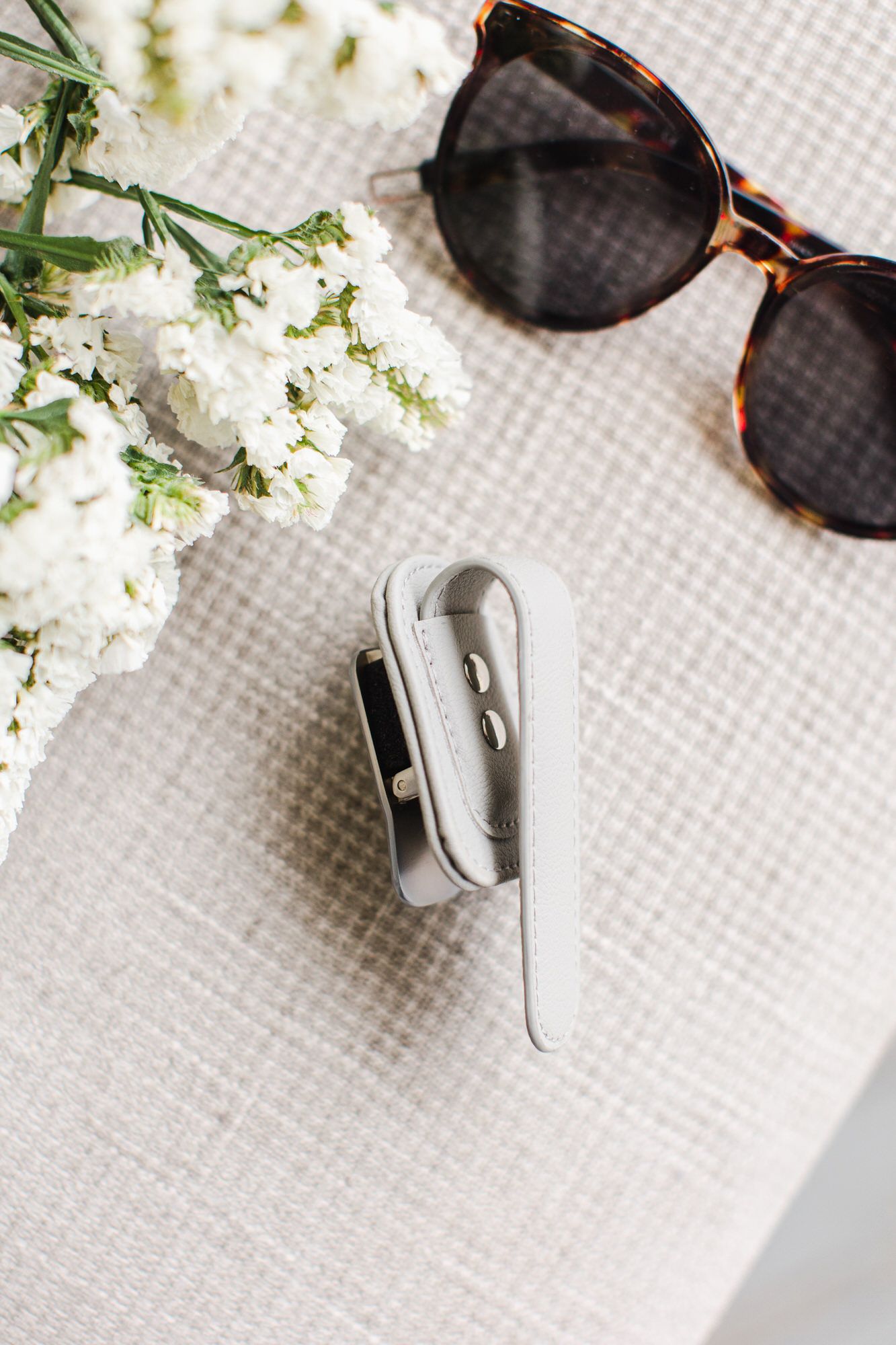 A pair of tortoiseshell sunglasses and white flowers laid on a light textured surface next to a silver paper clip.