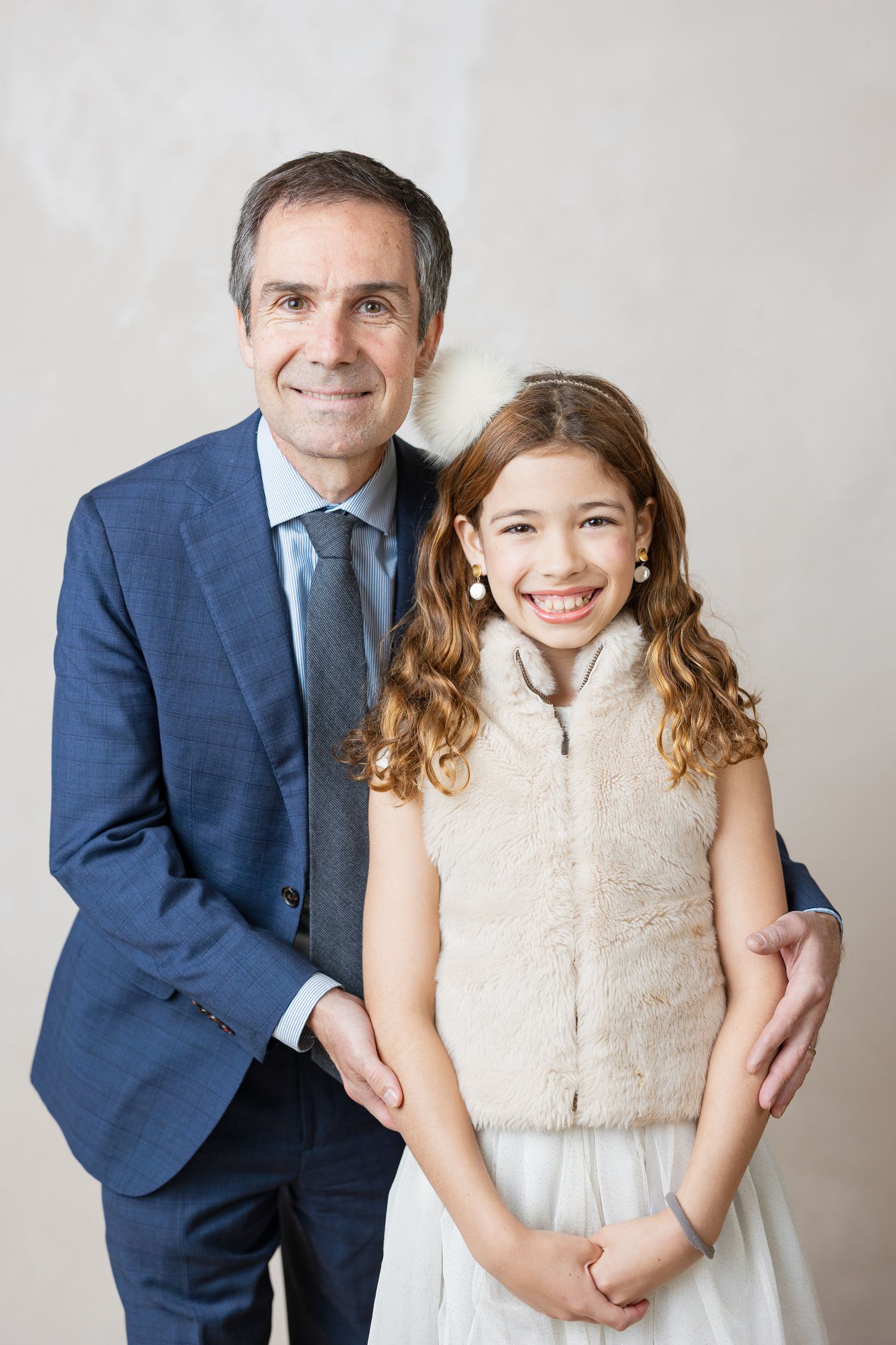 Portrait of a young girl in white dress and plush vest standing next to an adult in a navy suit and tie.