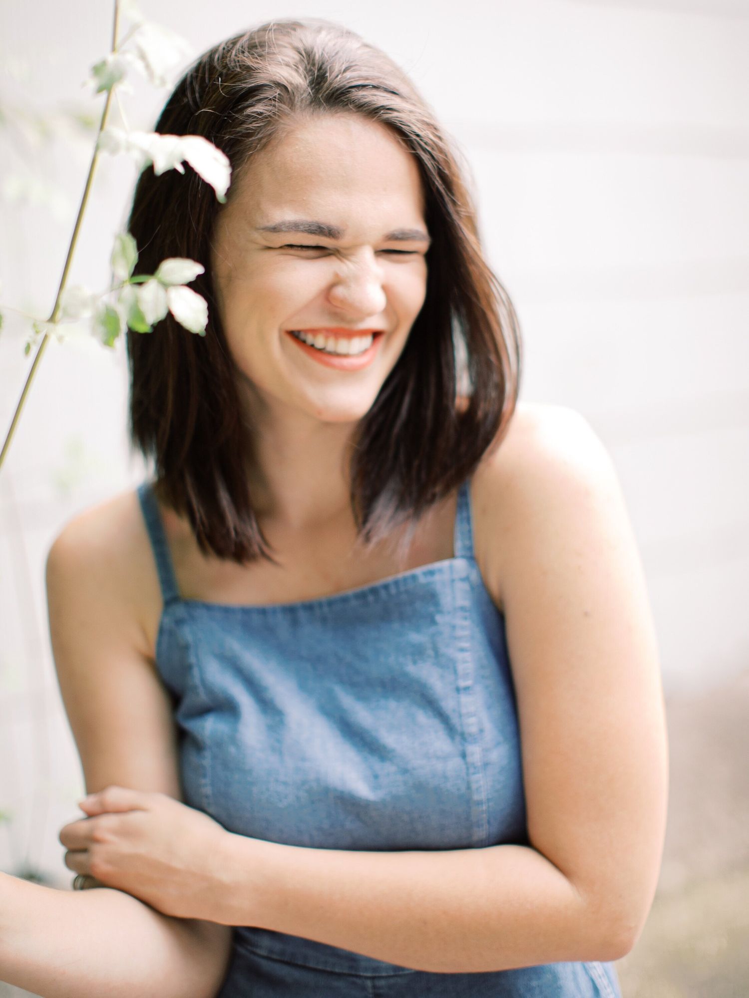 Joyful person in denim top laughs candidly while posing against light background with delicate white flowers nearby.