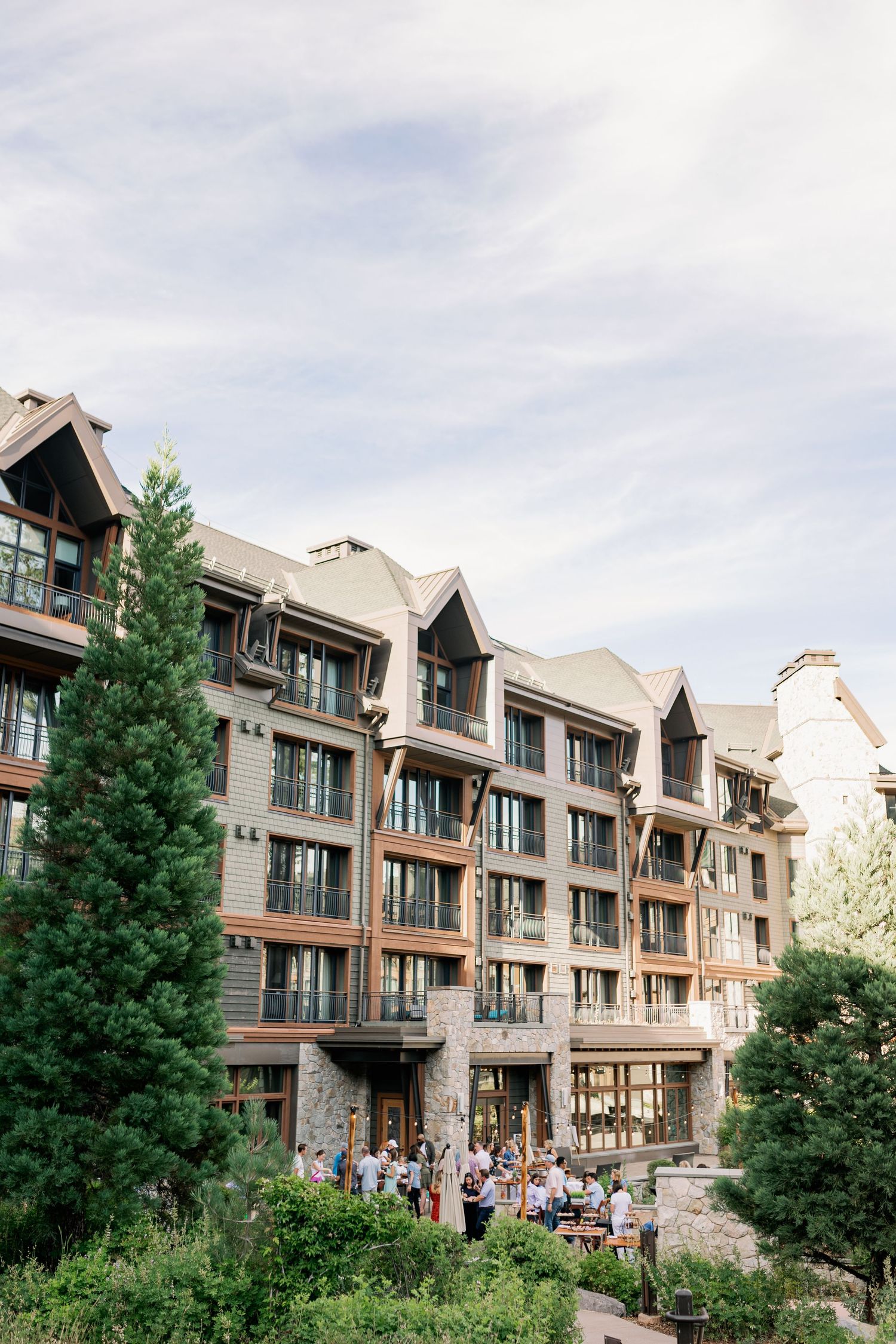 Upscale mountain resort building with multiple floors and balconies surrounded by evergreen trees against cloudy sky.