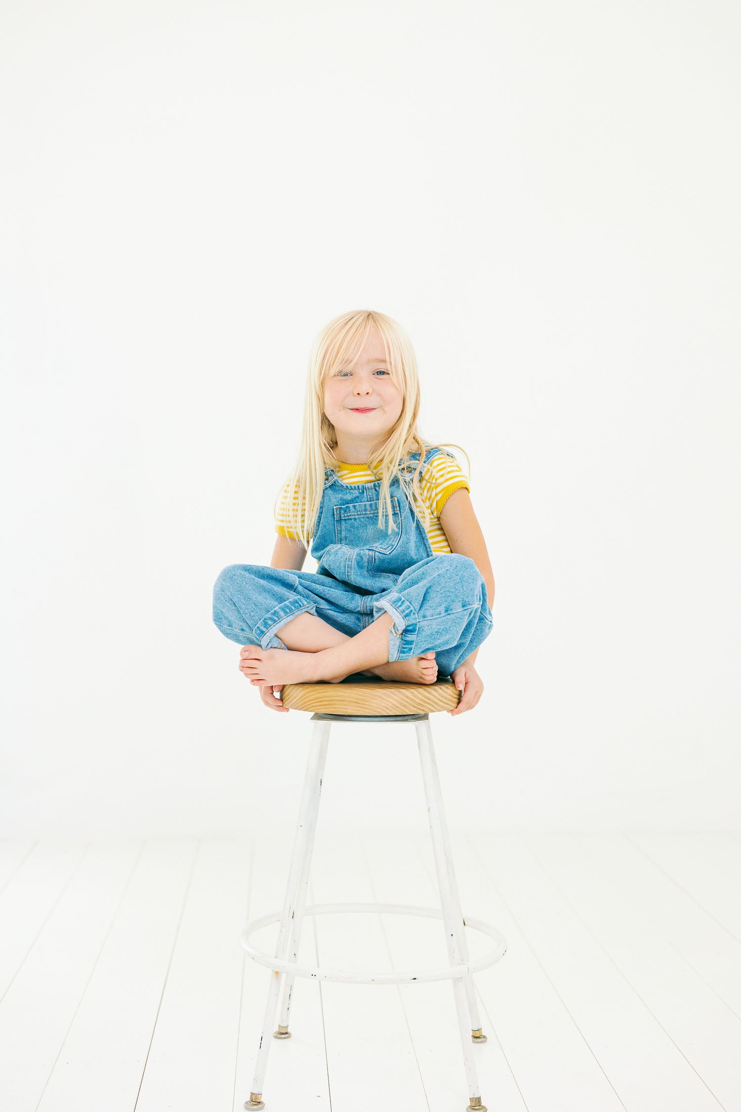 Child wearing blue overalls and yellow shirt sits cross-legged on white stool against white background.