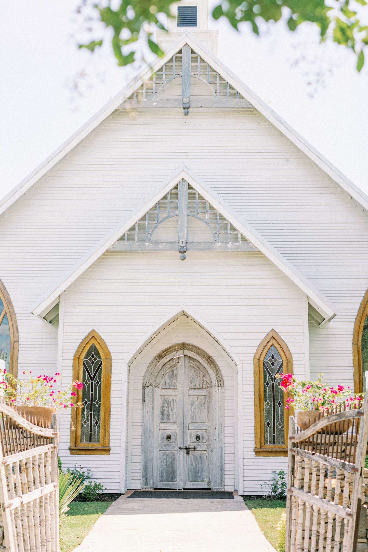 A charming white wooden chapel with pointed arch windows and door, decorated with pink flowers along the entrance path.