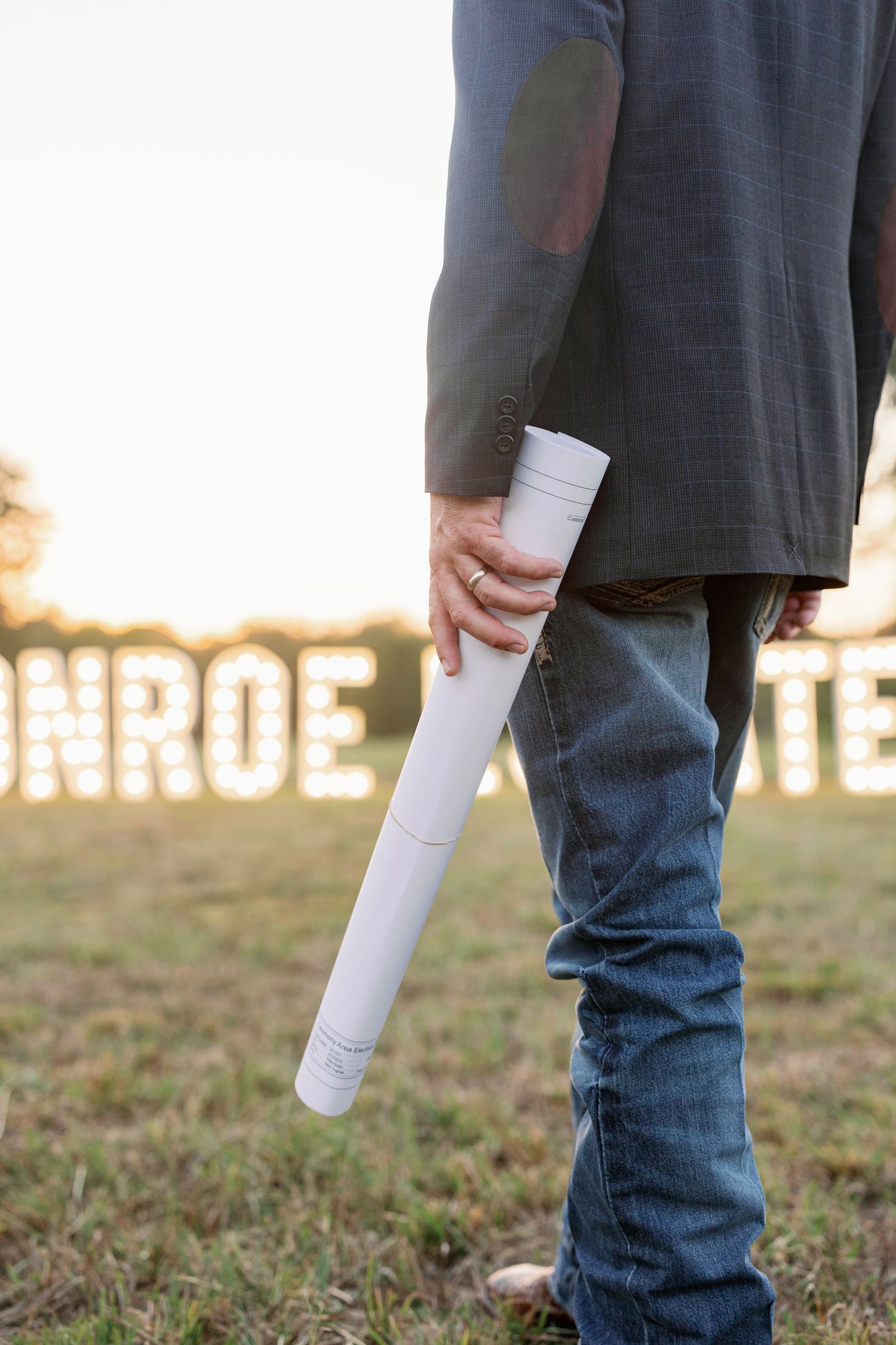 Person in dark suit and jeans holds white cylindrical tube outdoors at sunset with illuminated letters visible in background.