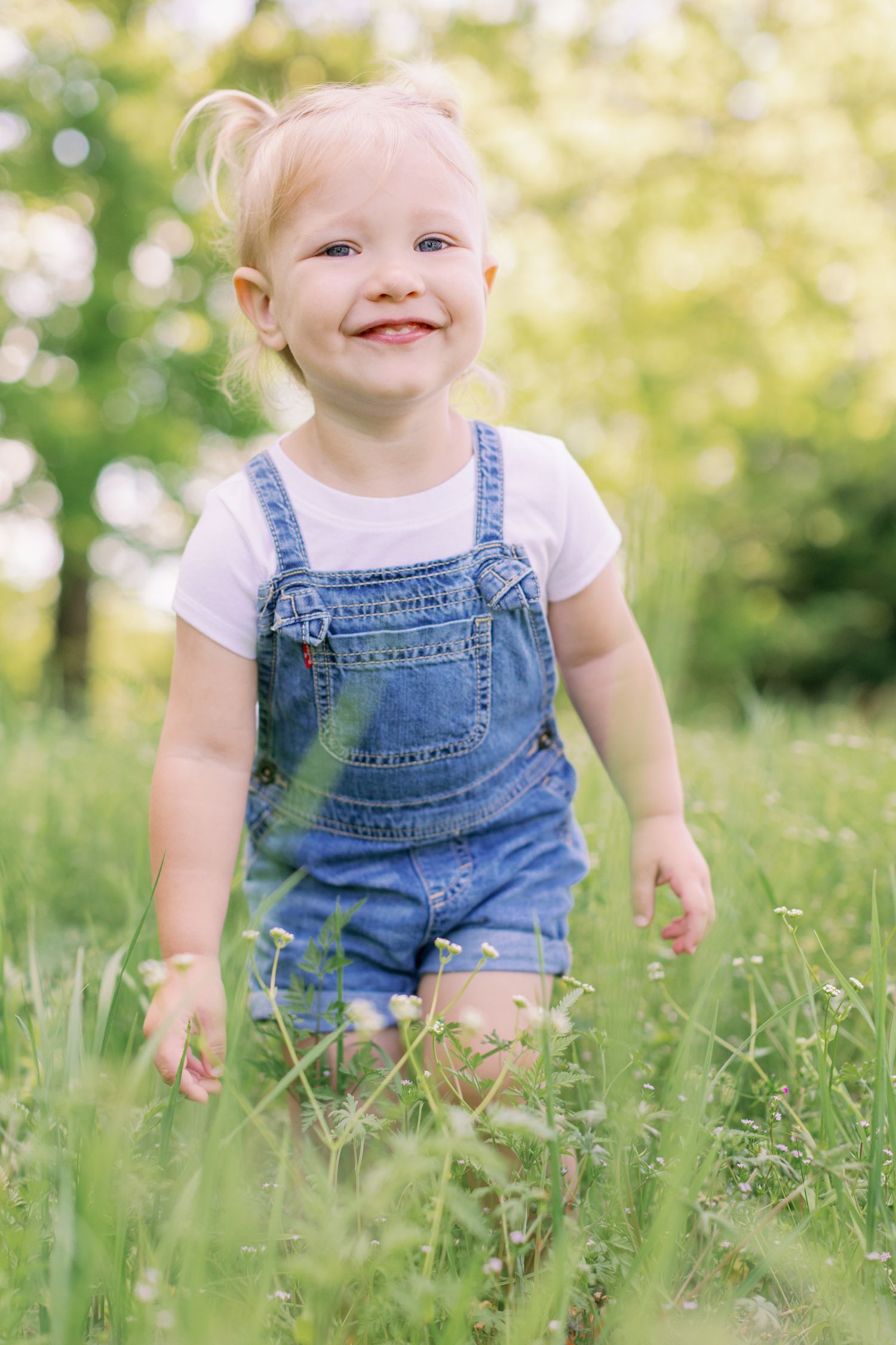 A happy child wearing denim overalls and a white t-shirt stands in a sunlit grassy meadow on a summer day.