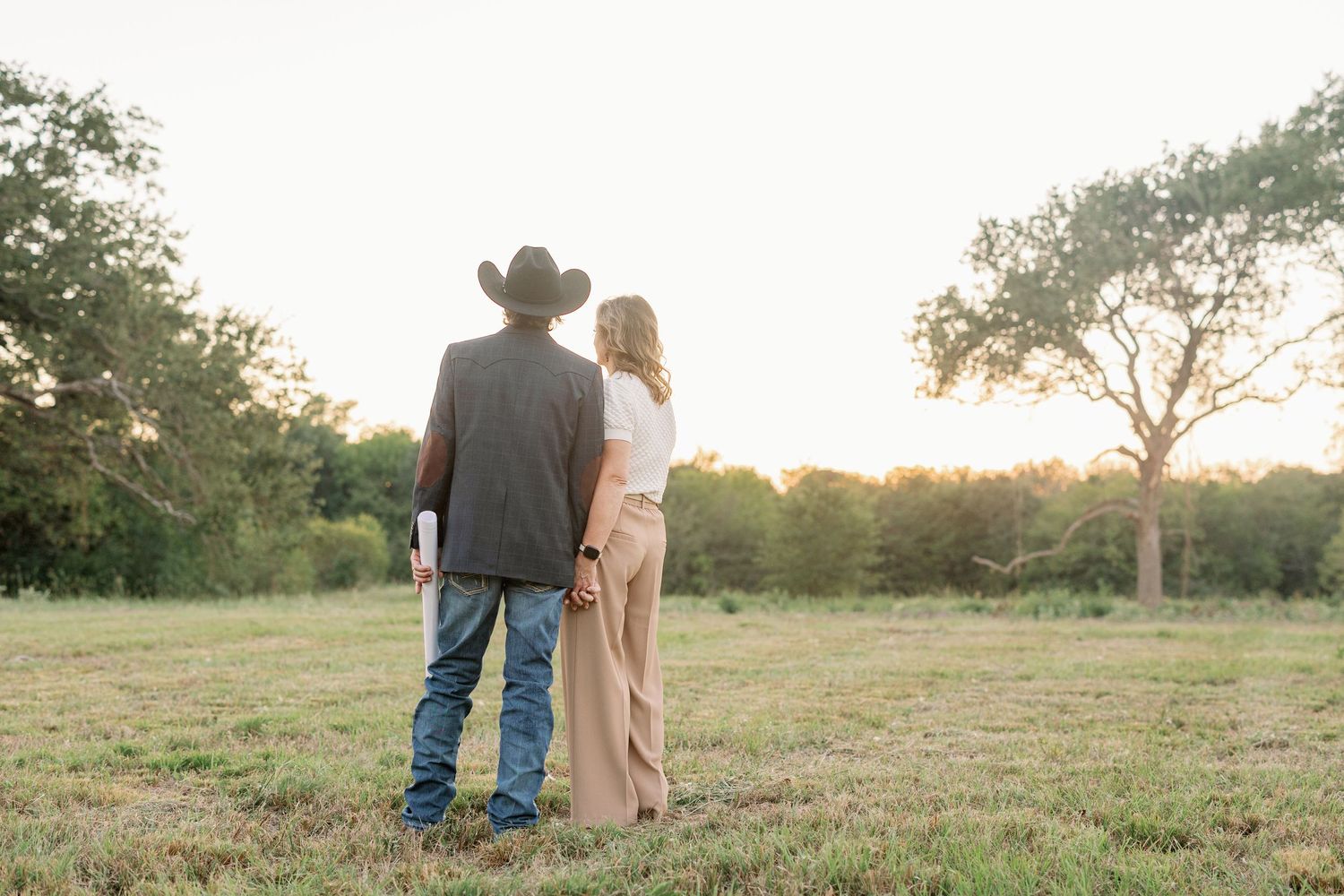 A couple stands together in a grassy field at sunset, with the person on the left wearing a cowboy hat and jeans.