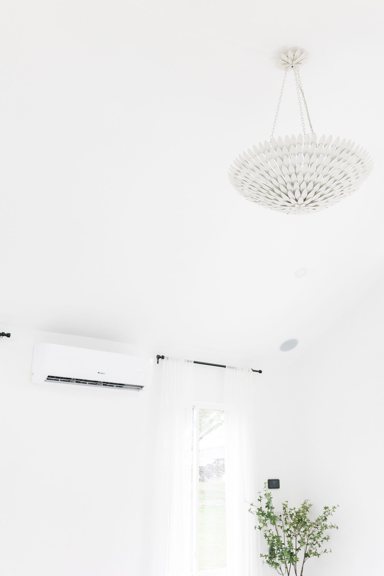 Modern white interior with elegant chandelier, sheer curtains, air conditioning unit, and potted plant against bright wall.
