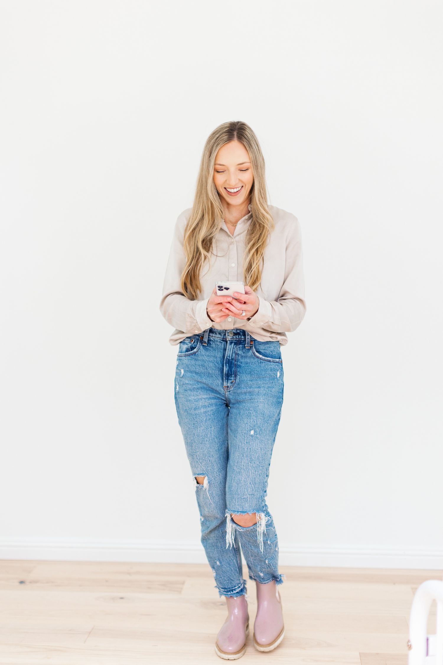 Woman in casual outfit of beige sweater and ripped jeans looks down at phone while smiling against white wall.